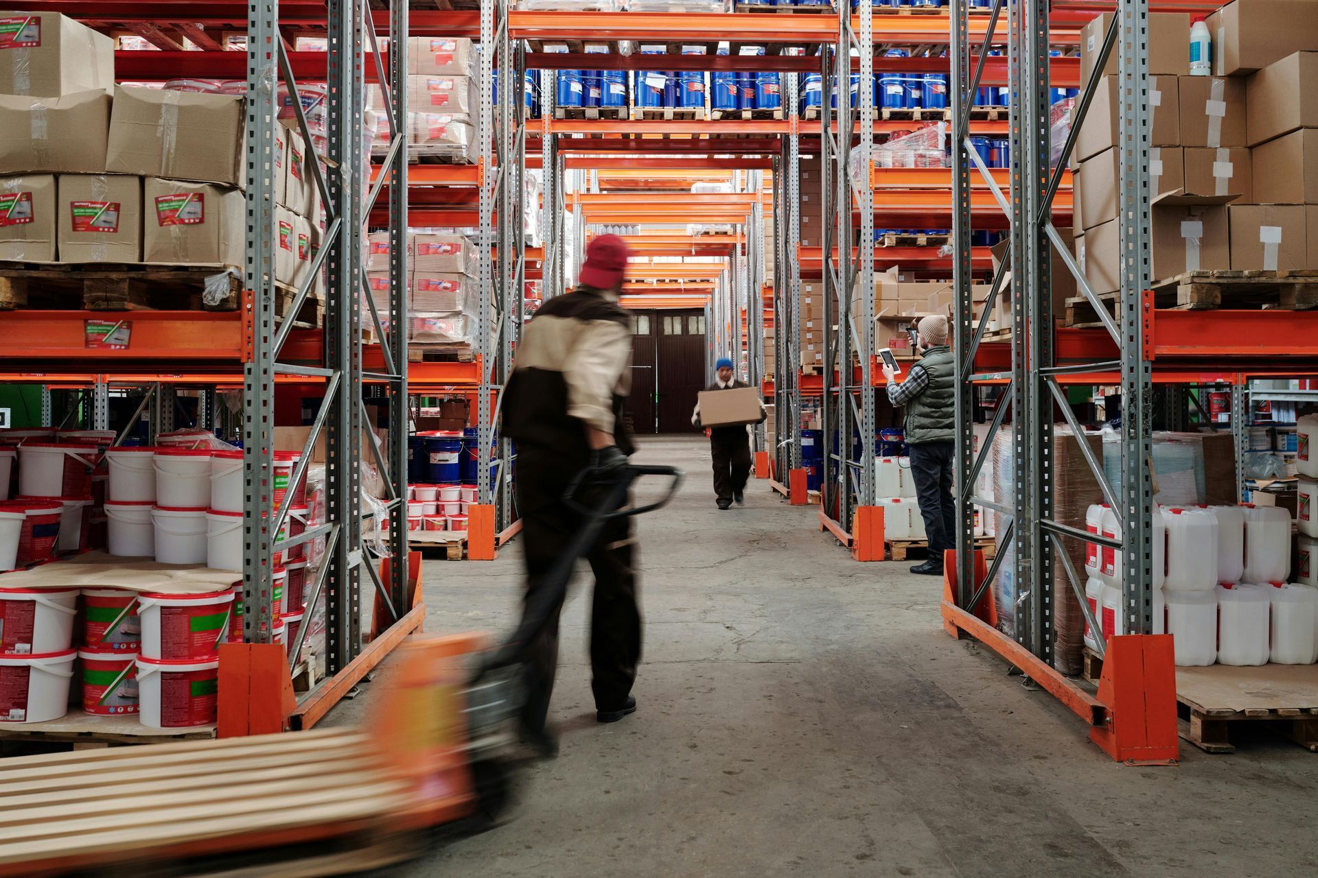 Warehouse workers moving goods; rows of shelves filled with boxes and materials.