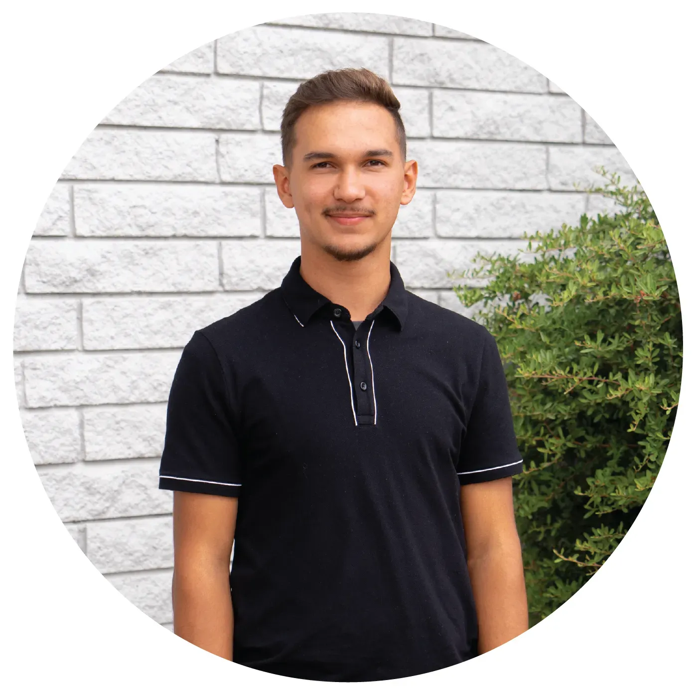A man in a black polo shirt is standing in front of a brick wall.