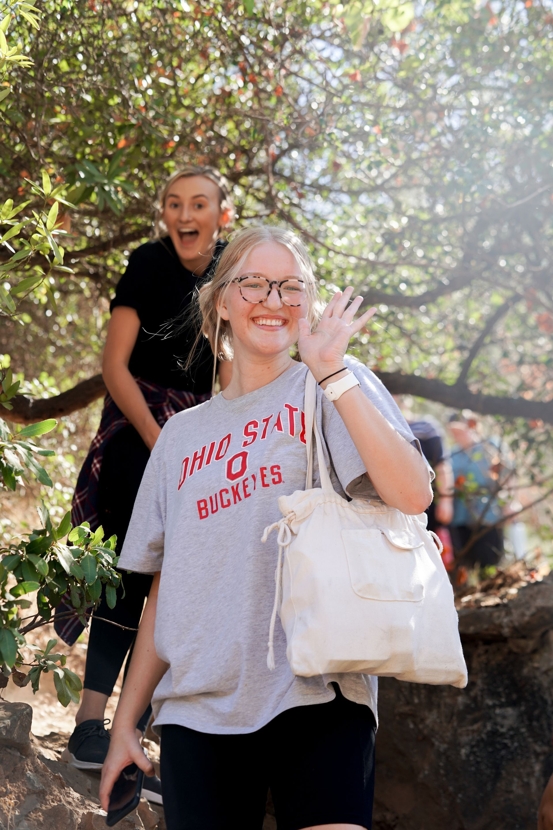 A girl in a gray shirt is holding a white purse and waving at the camera.