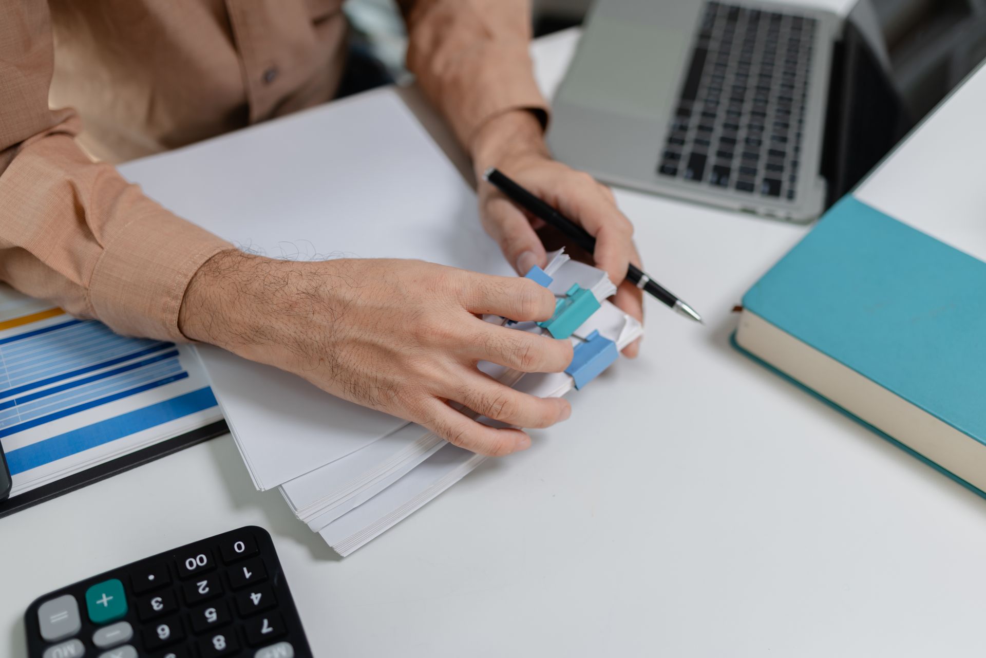 Hands holding a calculator and pen over paperwork beside a laptop and blue notebook