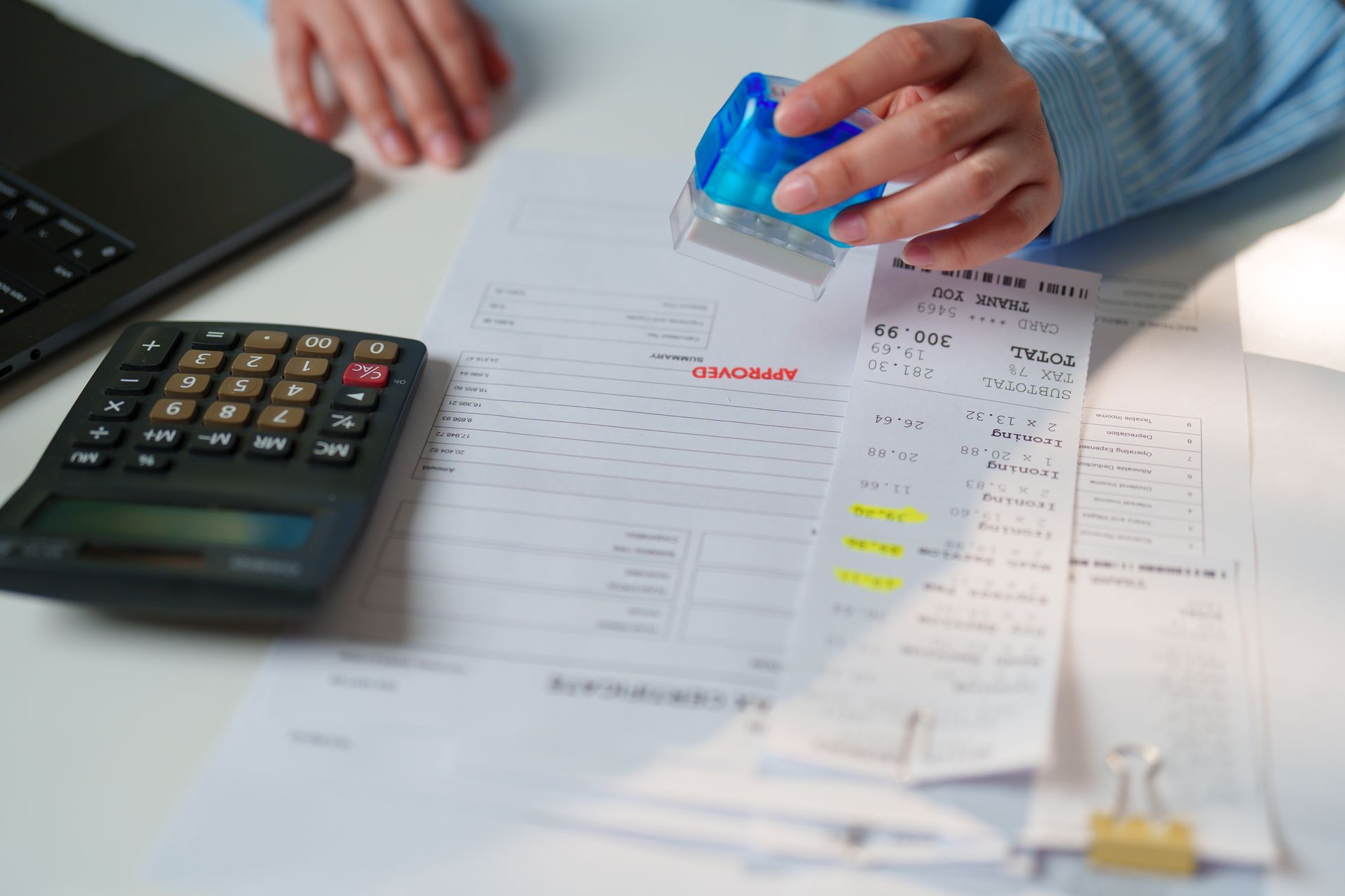 Hands stamping documents beside a calculator and laptop on a desk