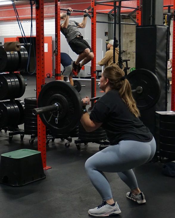 Woman squatting with barbell in a gym, man doing pull-ups in background.