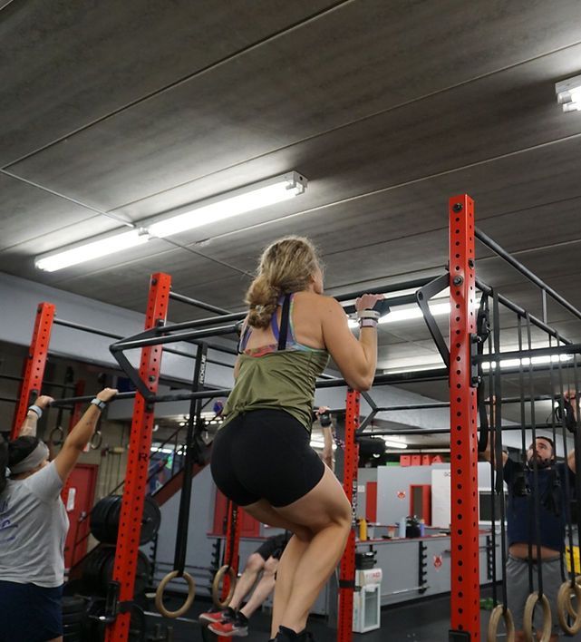 Woman doing a pull-up at a gym. She's wearing black shorts and a green tank top, with others nearby using equipment.