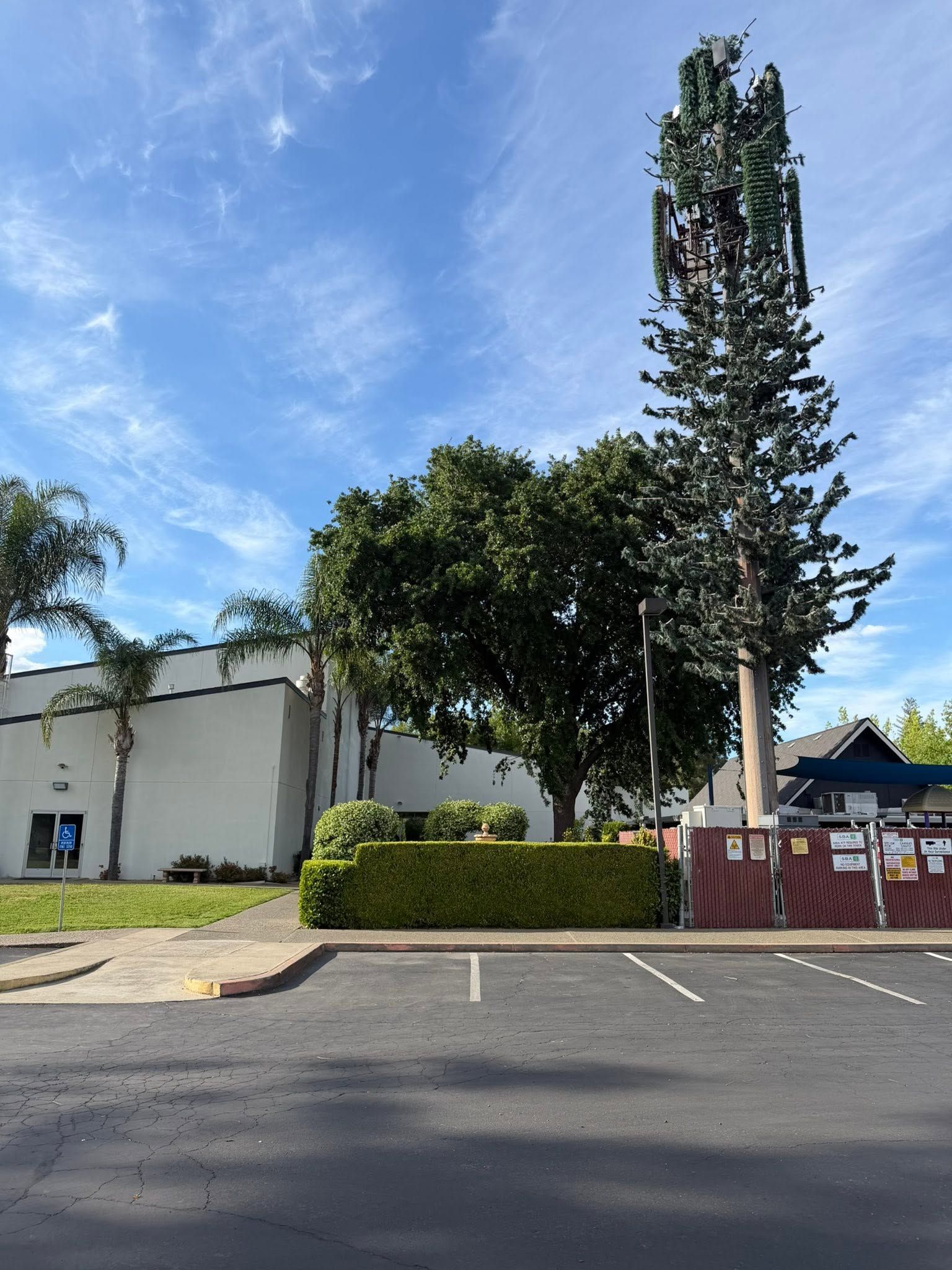 Parking lot beside a white building, with a tree, fenced area, and blue sky.