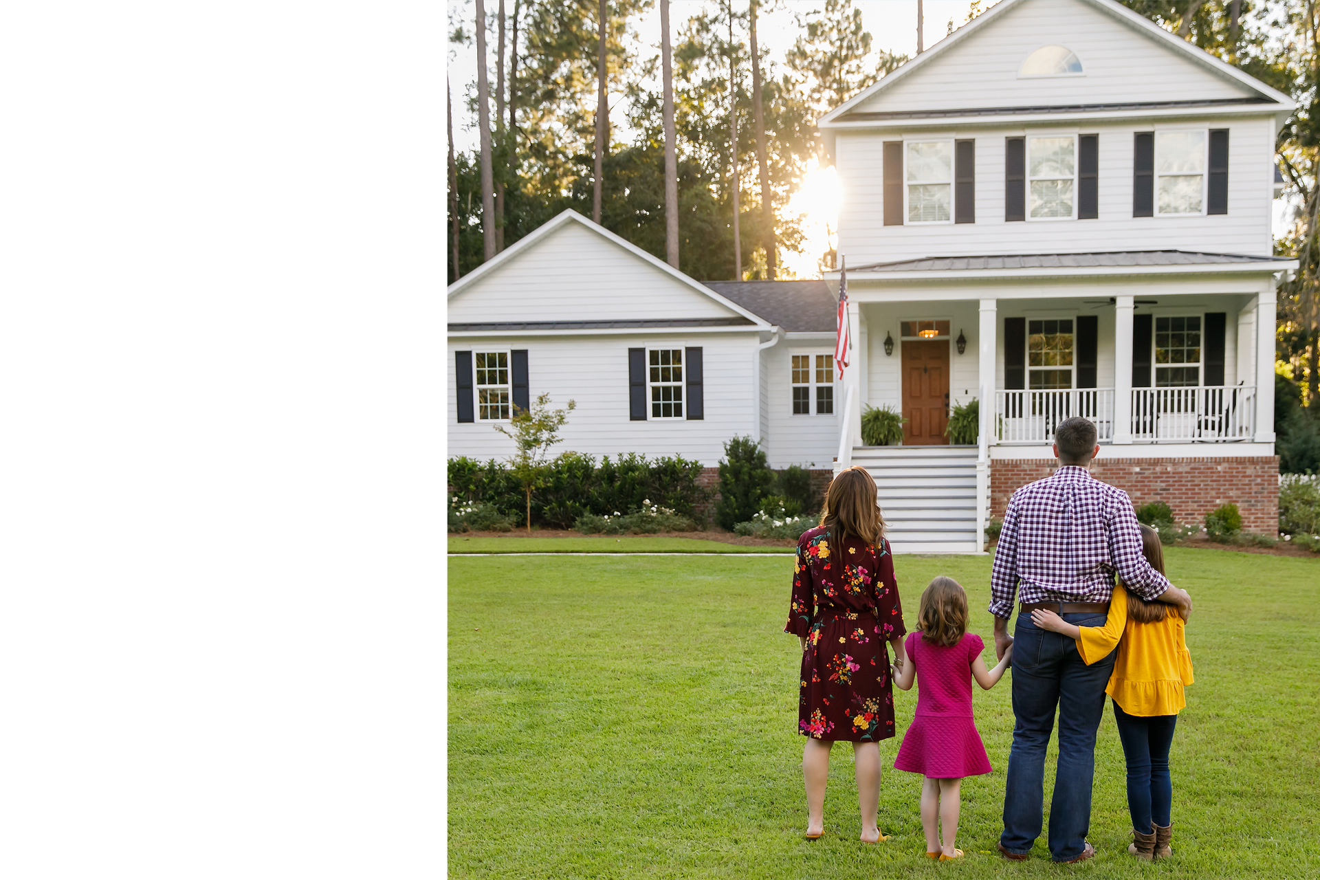 Family of four standing on a lawn facing a white house with a porch and lit windows at dusk