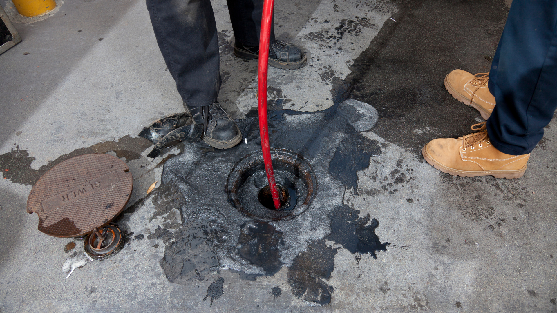 Workers clearing a clogged manhole with a red hose on a wet street near a storm drain