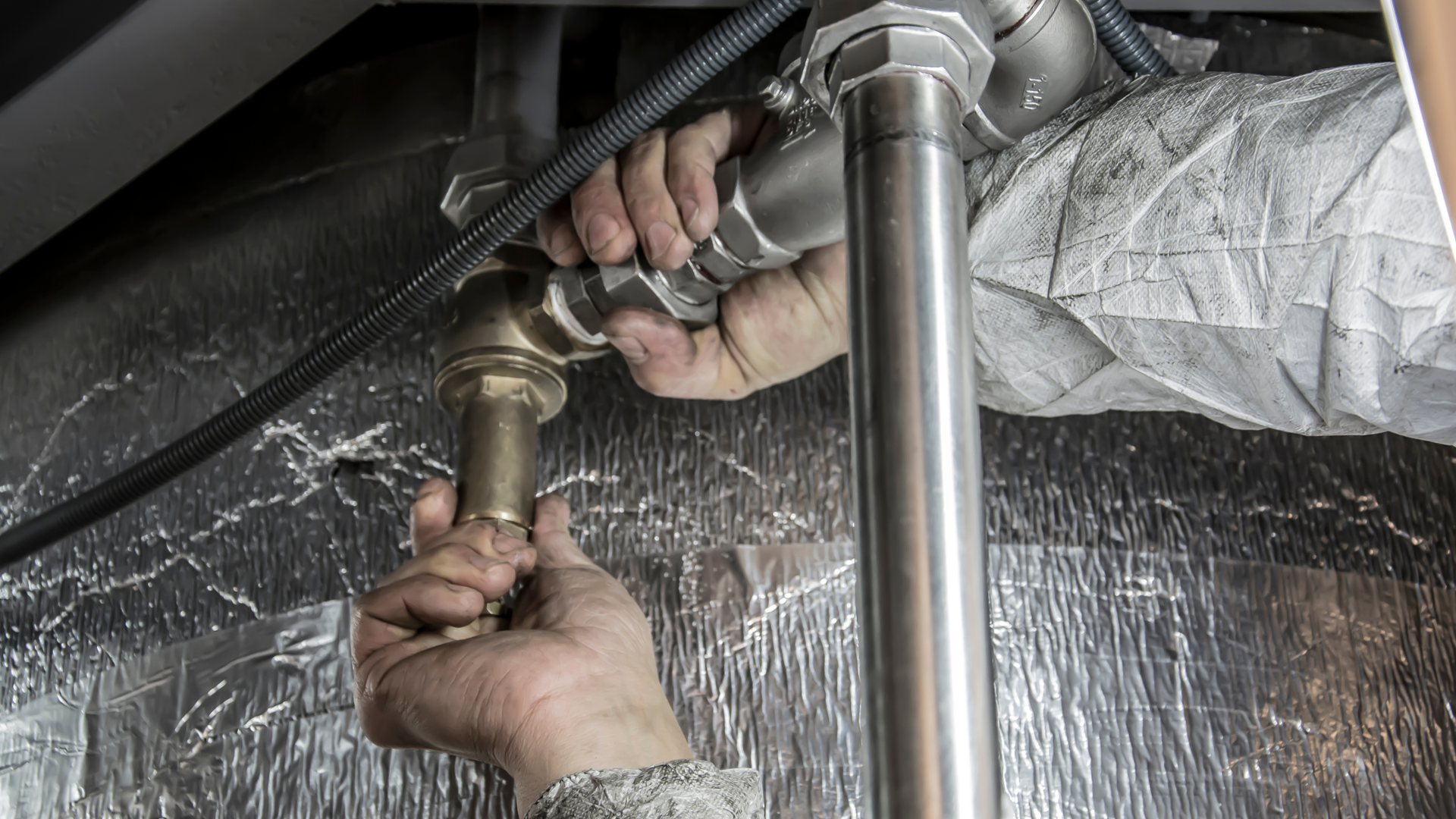 Hands holding a hose nozzle spraying water under a sink against a plastic-covered wall.
