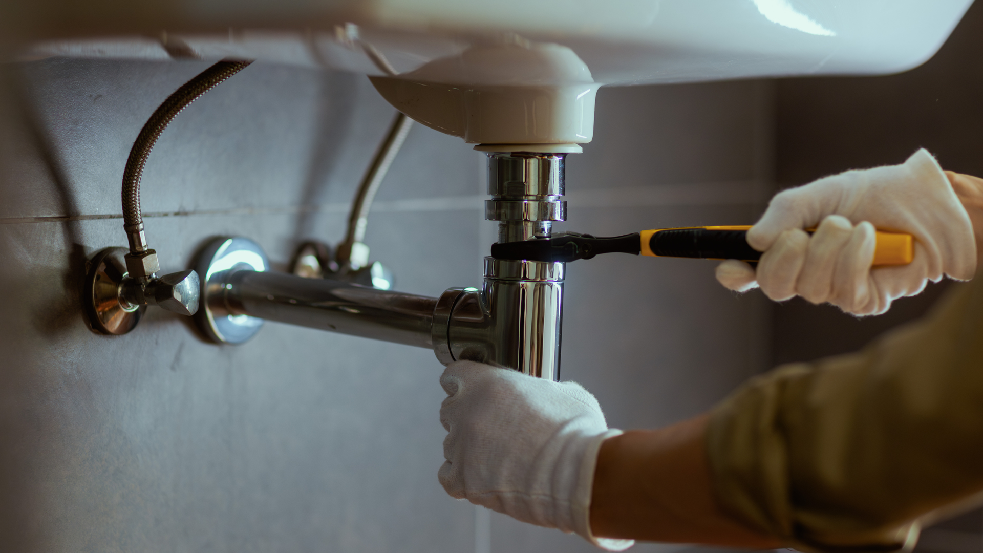 Gloved hands tightening a chrome pipe under a sink with a screwdriver