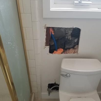 Bathroom wall damaged behind toilet, exposing pipes and rough drywall near a window.