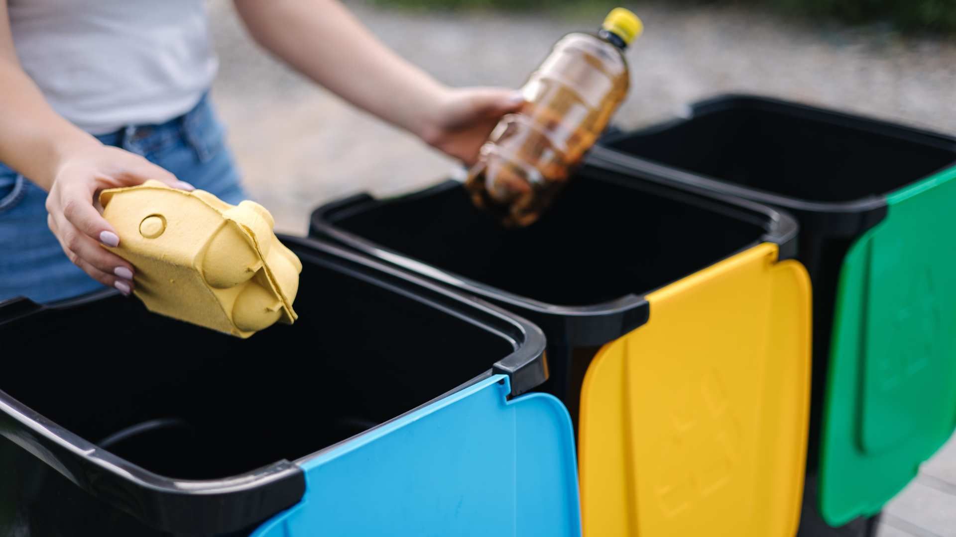 Hands sorting recyclables into blue, yellow, and green bins outdoors