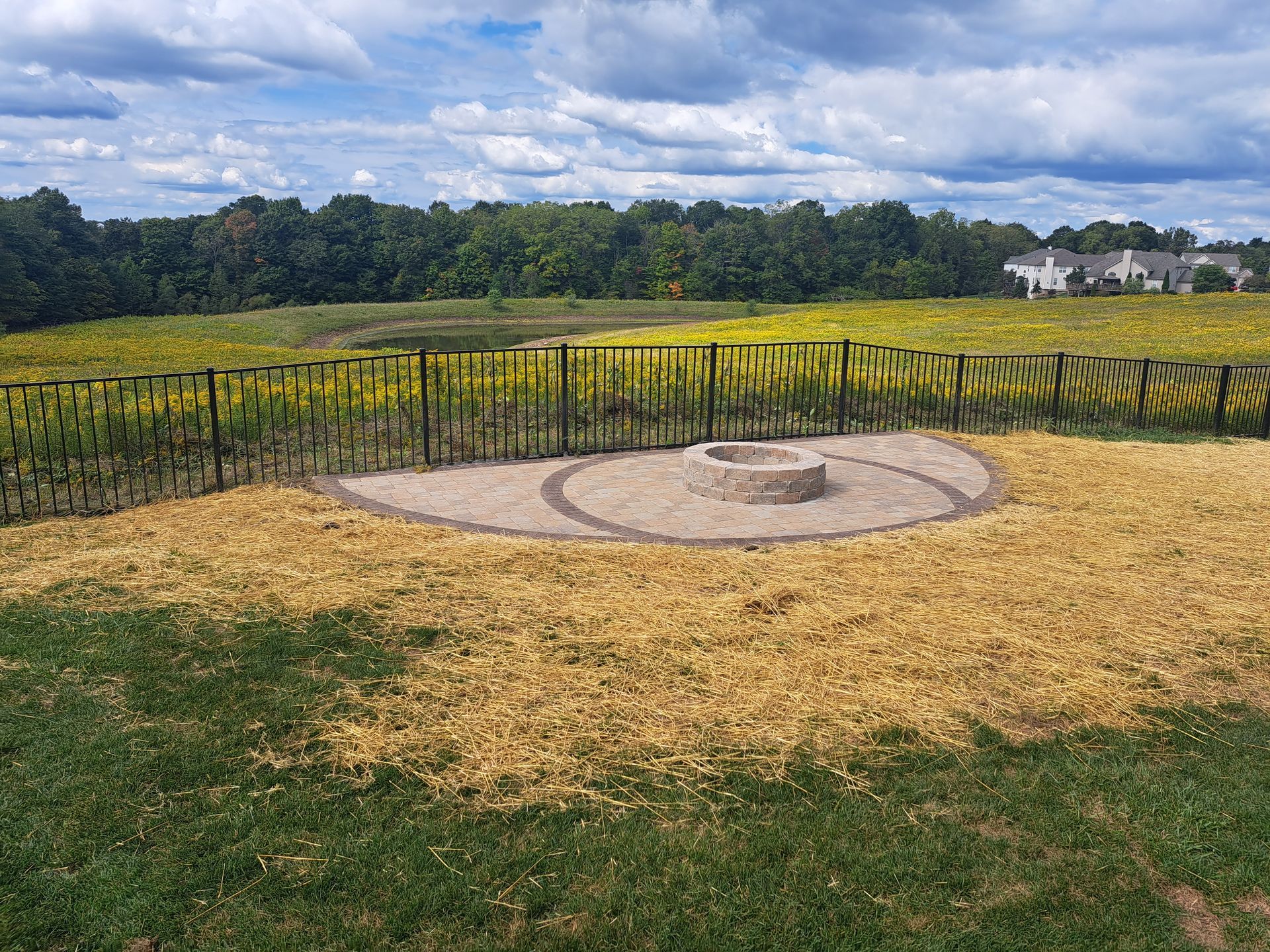 Fire pit area with stone circle, surrounded by tall grass and a metal fence, in a field under a cloudy sky.