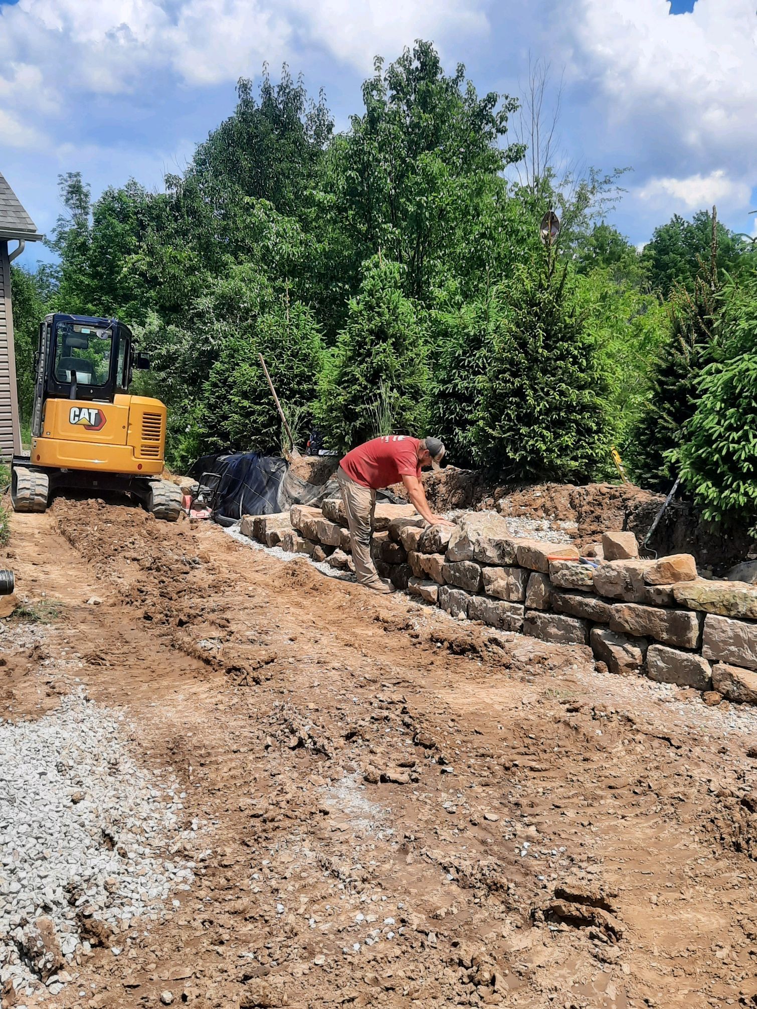 Man building a stone retaining wall with a small yellow excavator nearby, outdoors.