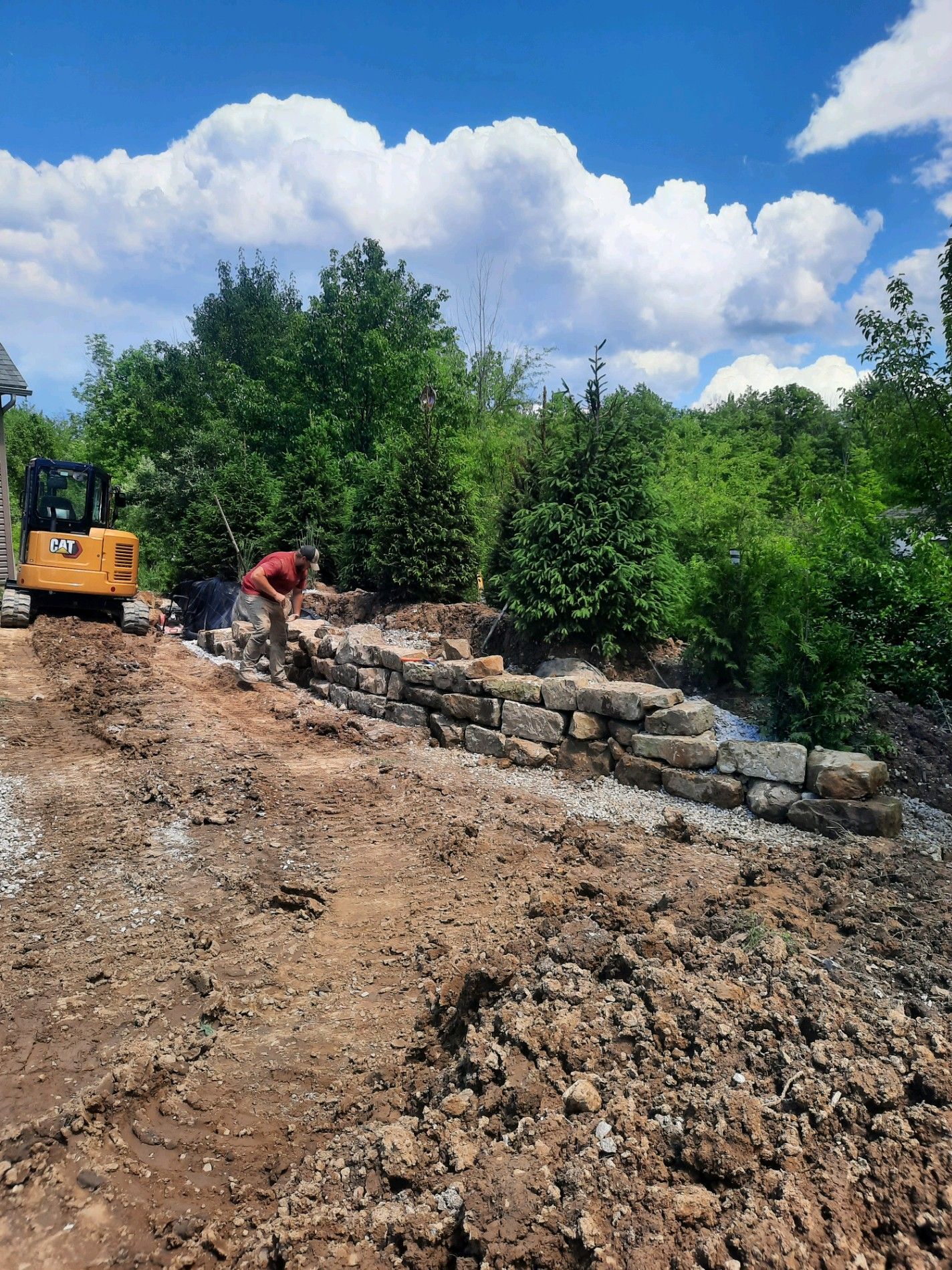 Man working on stone wall construction with a small excavator, outdoors on a sunny day.