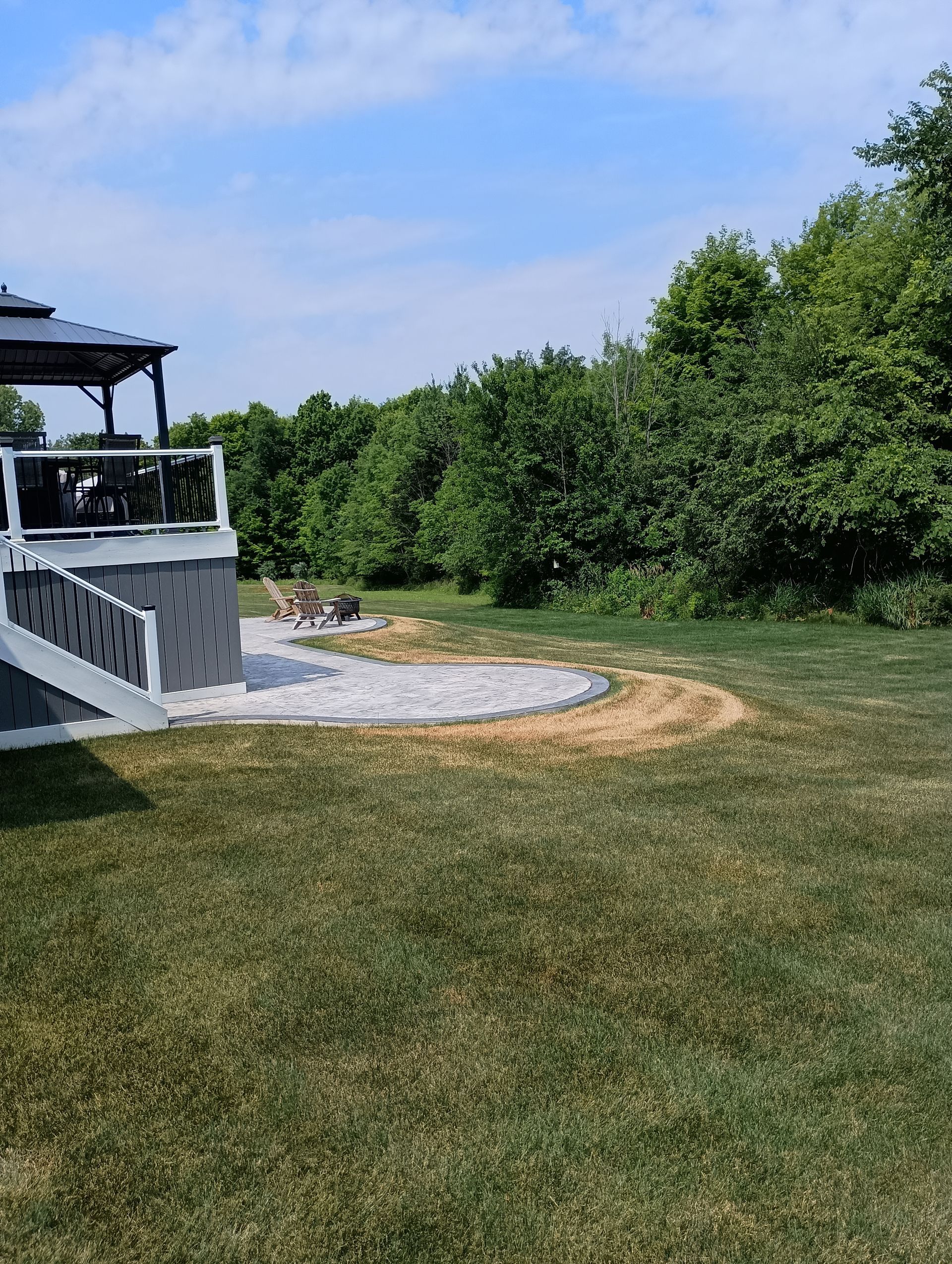Backyard with a deck, a circular stone patio, and green grass leading to trees under a blue sky.
