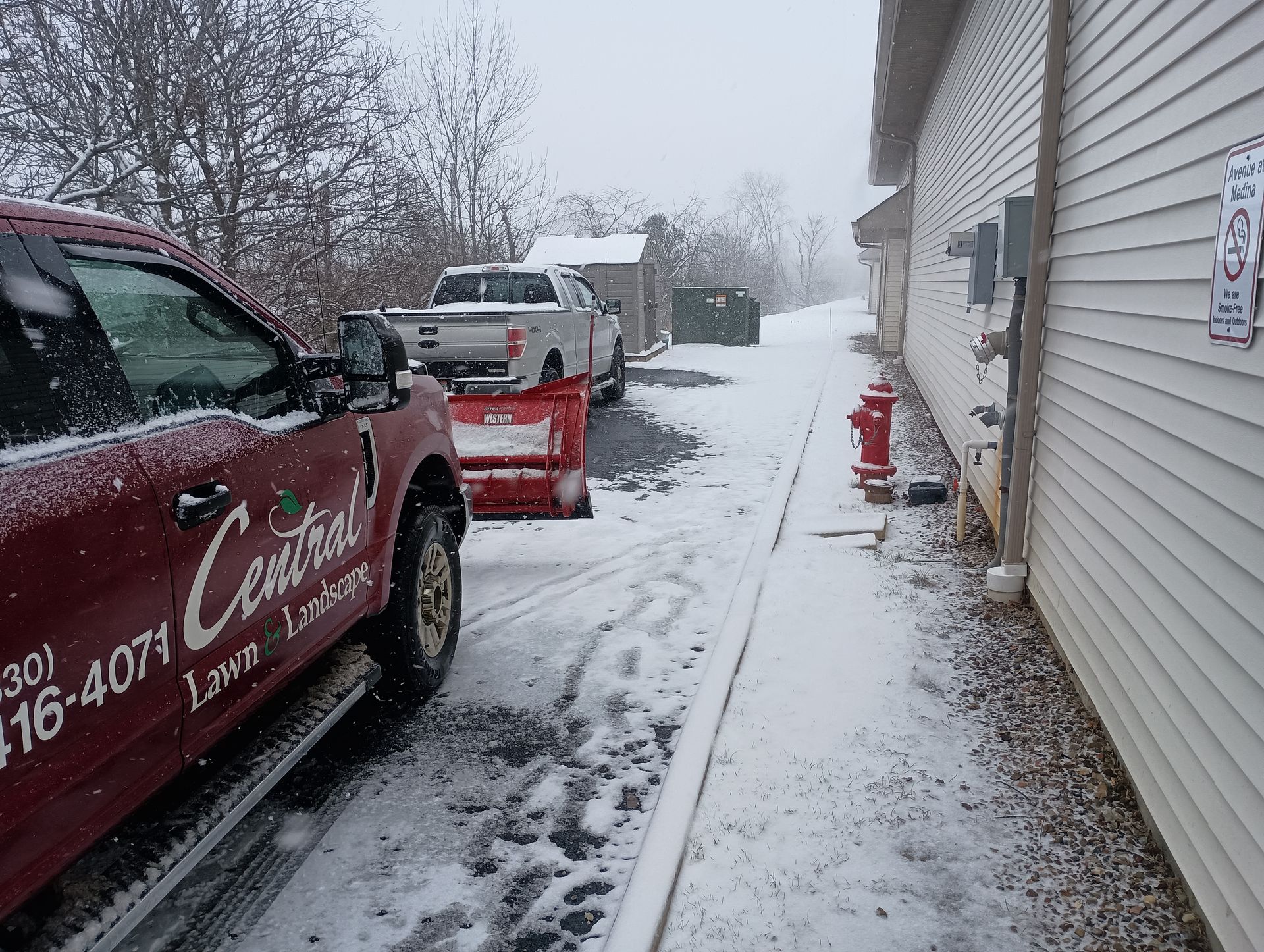 Snowy scene with red Central truck plowing driveway next to building. Another truck with snow plow.