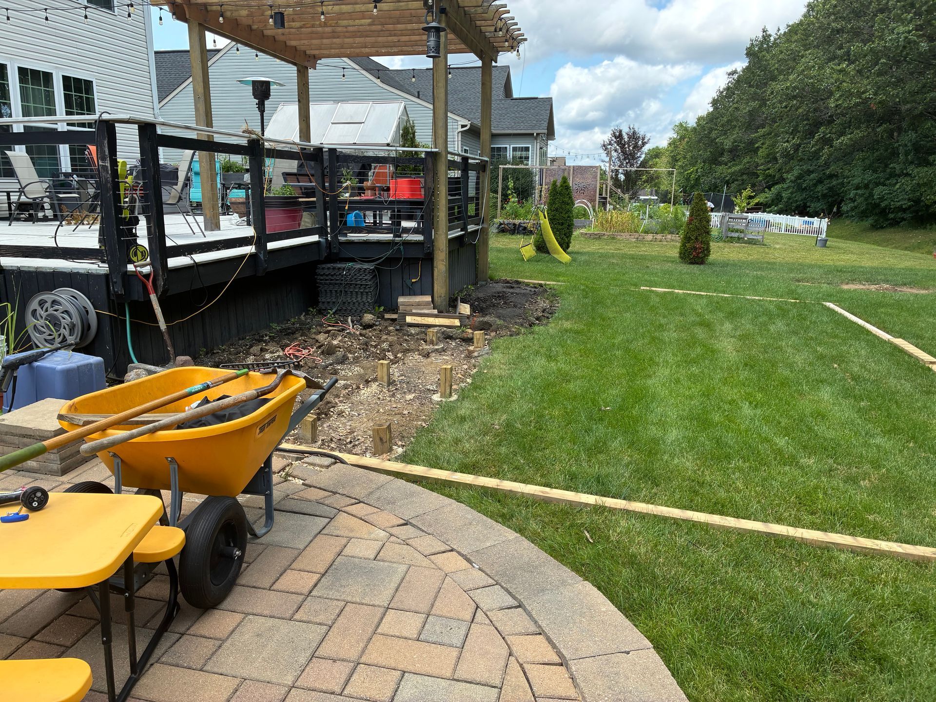 Backyard with a deck, patio, and lawn; wheelbarrow and construction materials visible.