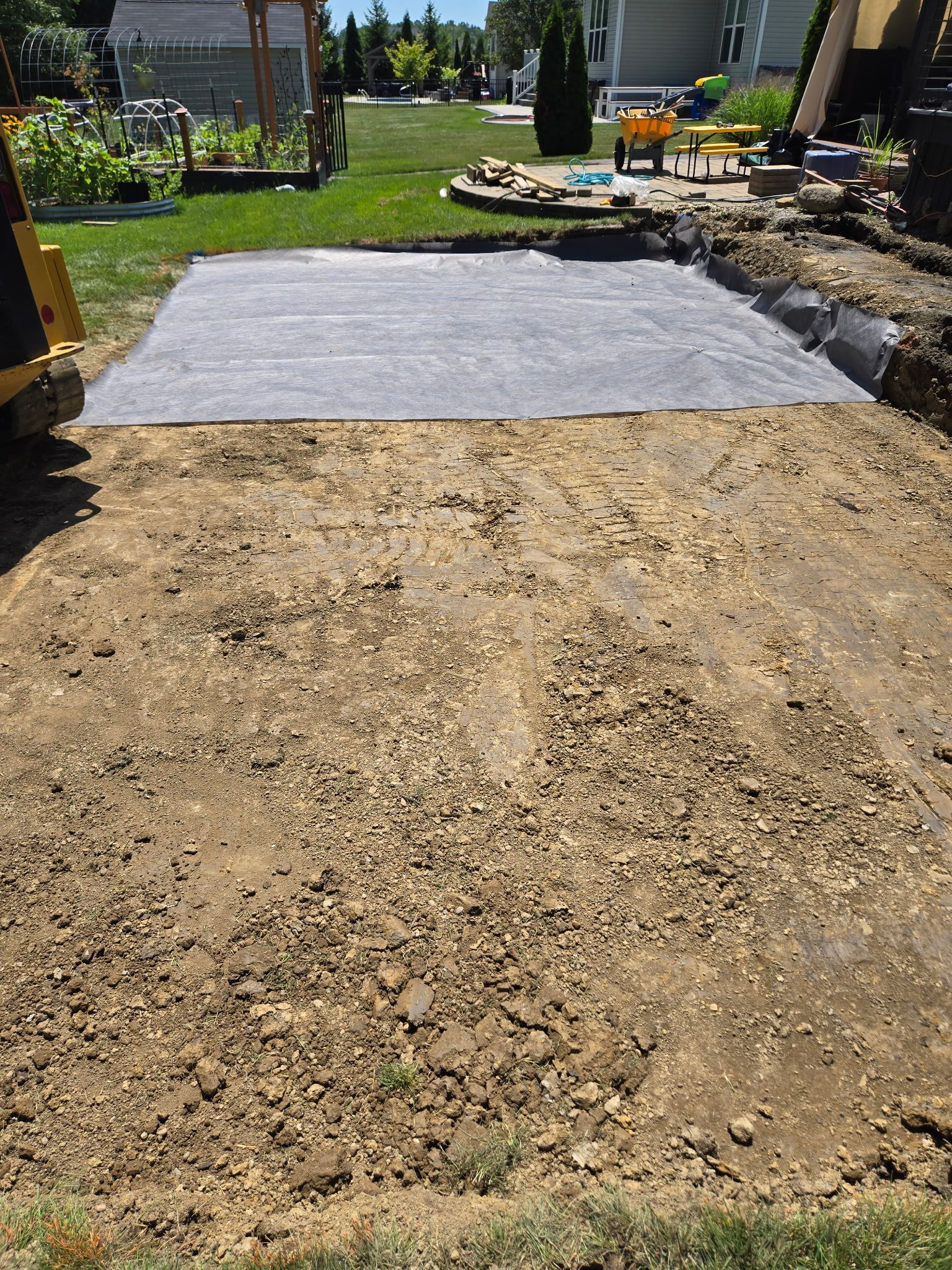 Excavated yard with dark fabric laid, possibly for a patio. Soil visible, construction equipment in view.