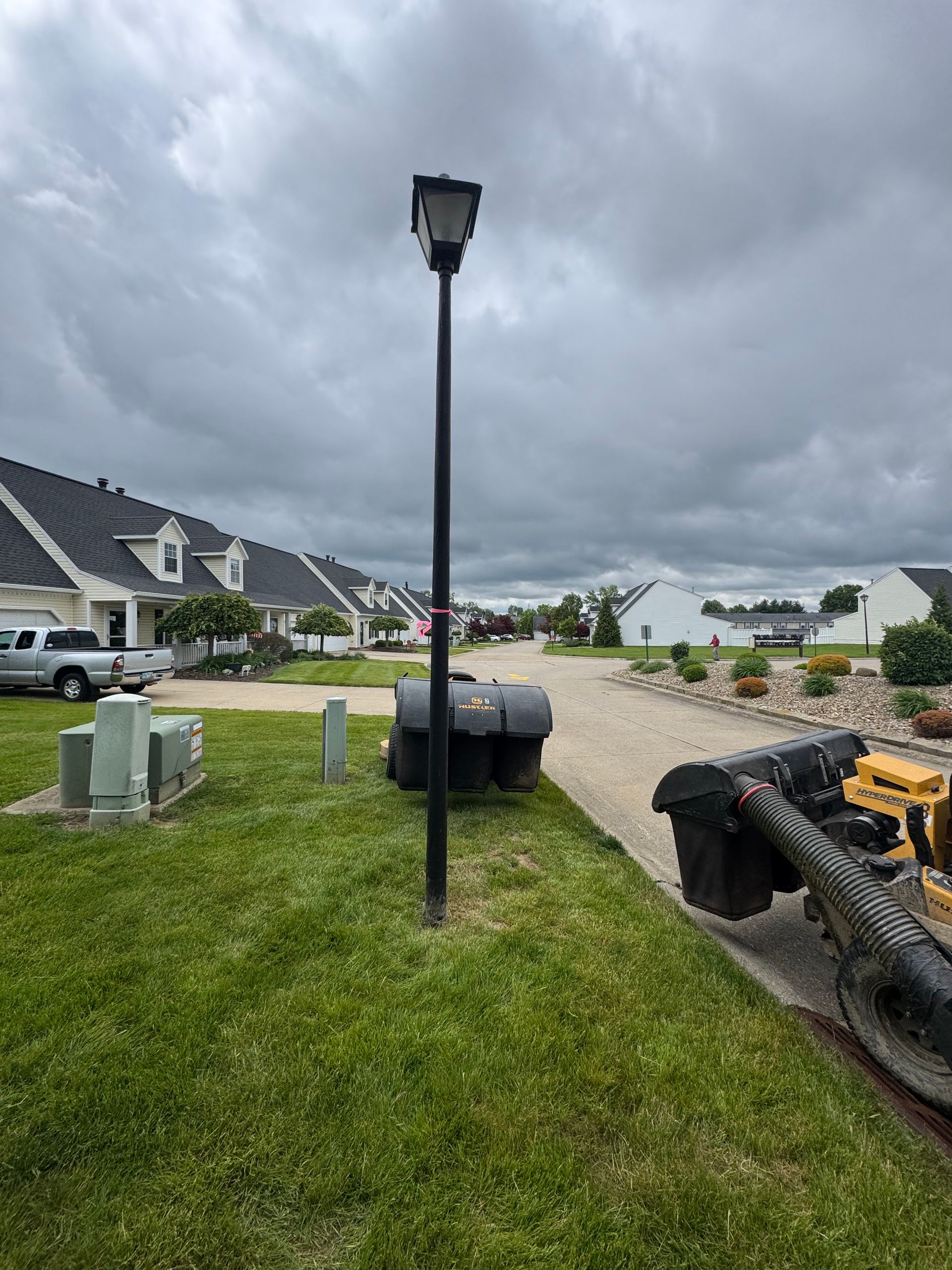 A black street light stands on green grass. A large machine and houses are in the background under a cloudy sky.