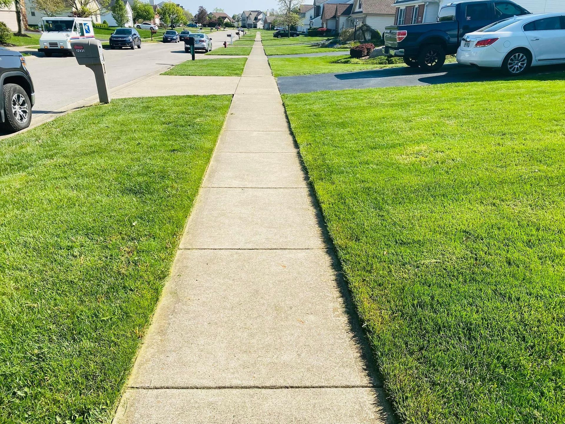 Sidewalk running straight ahead, bordered by green grass and houses in the background. Sunny day.