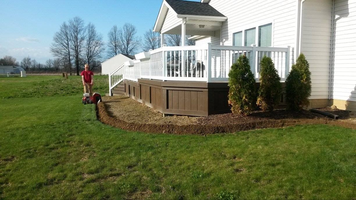 Person mowing grass near a house with a deck and landscaping; sunny day.
