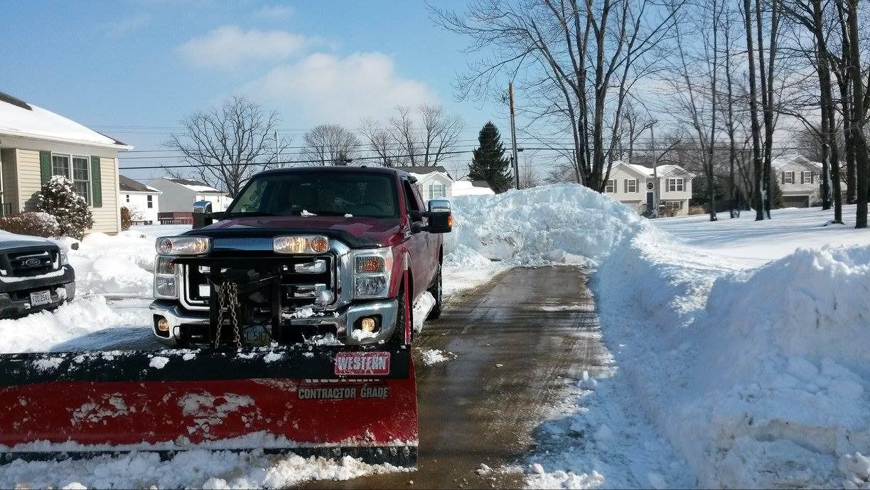 Red snow plow truck clearing a snow-covered street on a sunny day, with houses in the background.