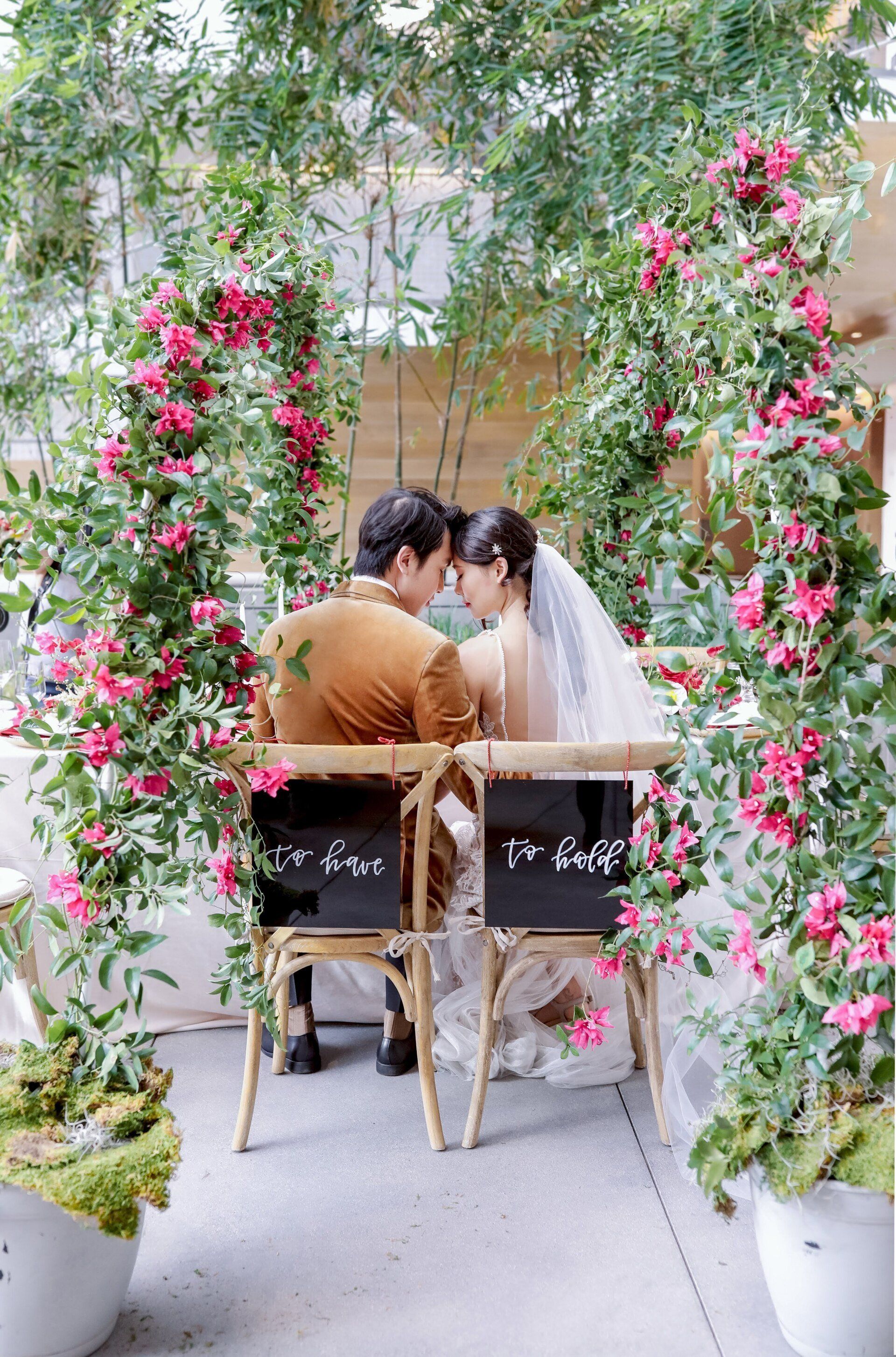 A bride and groom are sitting in chairs under a flower arch.