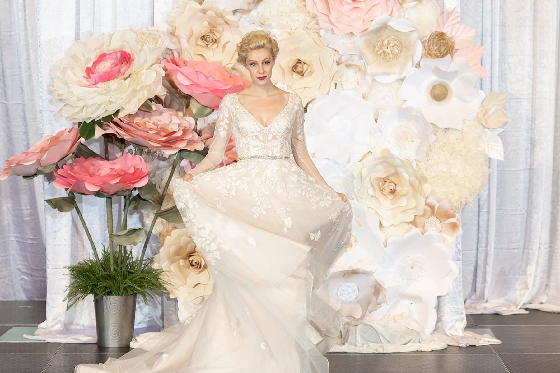 A woman in a wedding dress is standing in front of a wall of paper flowers.