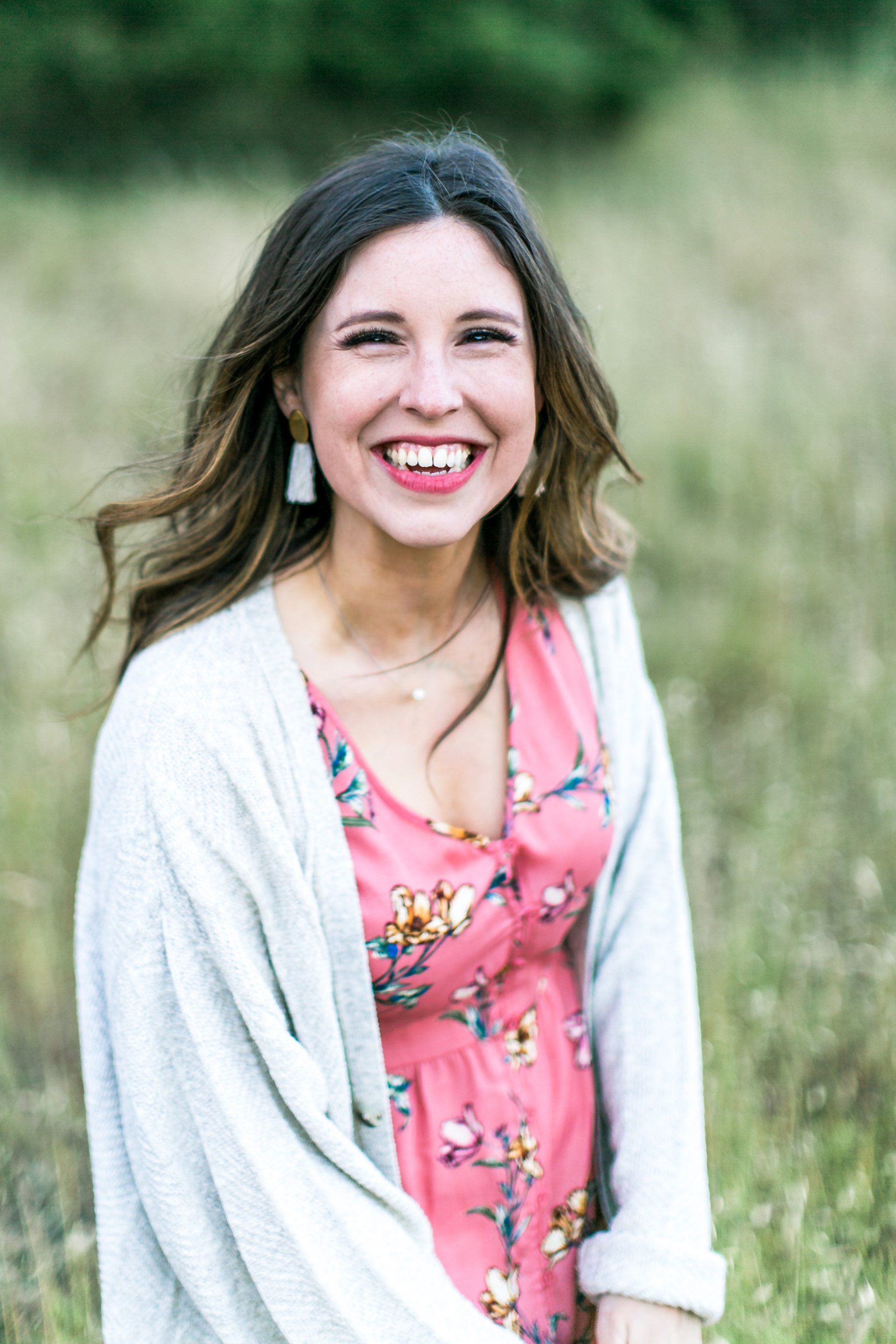 A woman in a pink dress and gray cardigan is smiling in a field.