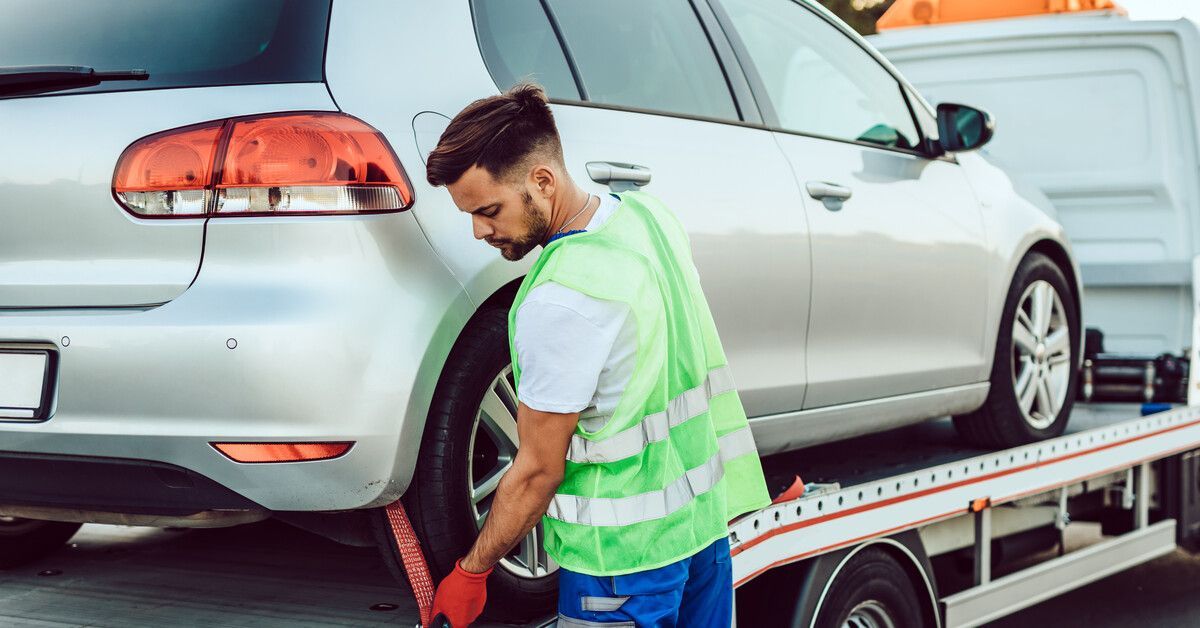 A man in a green safety vest using a harness to secure a silver vehicle to the trailer of a truck.