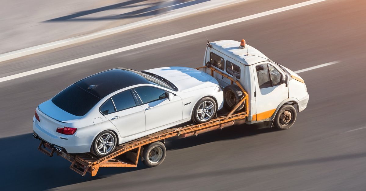 An overhead view of a truck transporting a white car on a flatbed down an open multi-lane road.