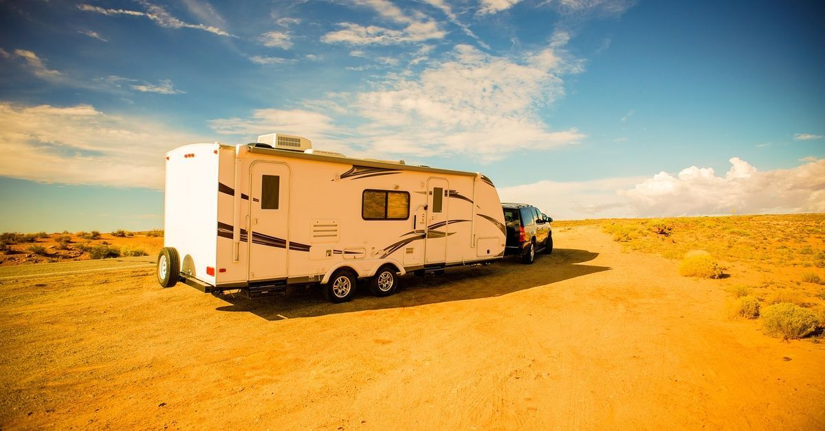 A black SUV towing a large white RV on an orange dirt road in the desert under a bright, sunny sky.