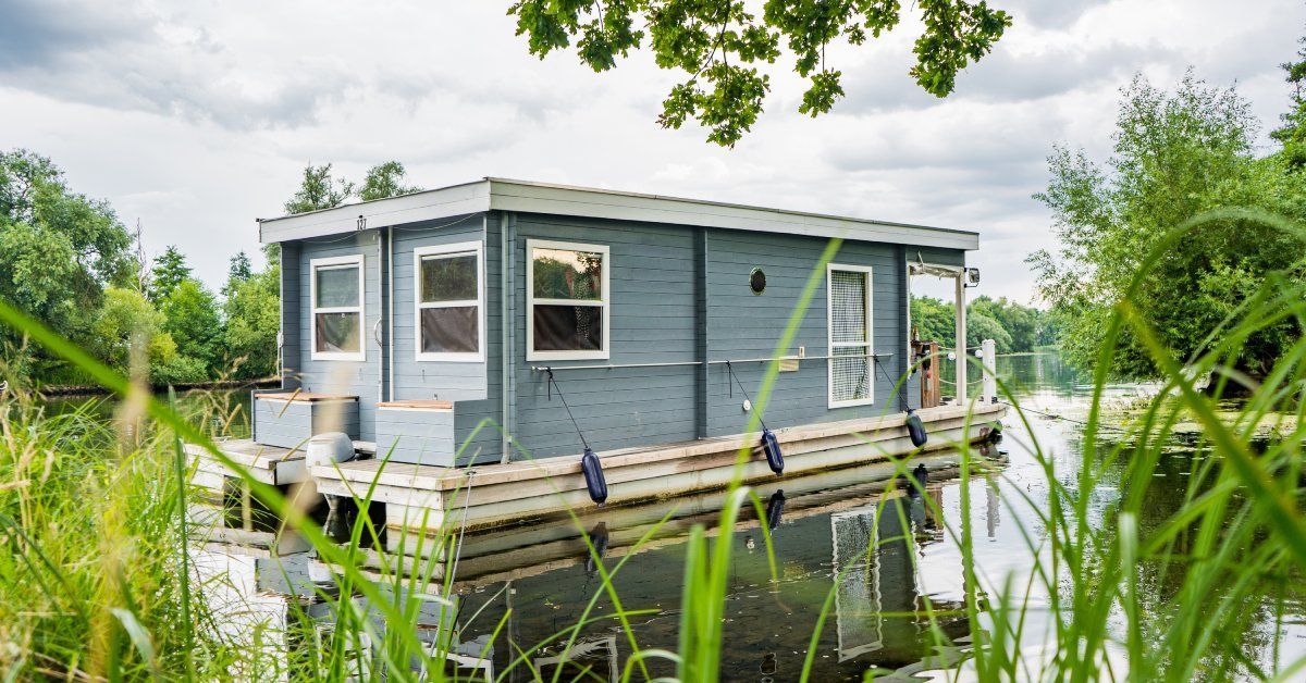 A blue and white house boat floating in a body of water surrounded by trees and tall grasses.
