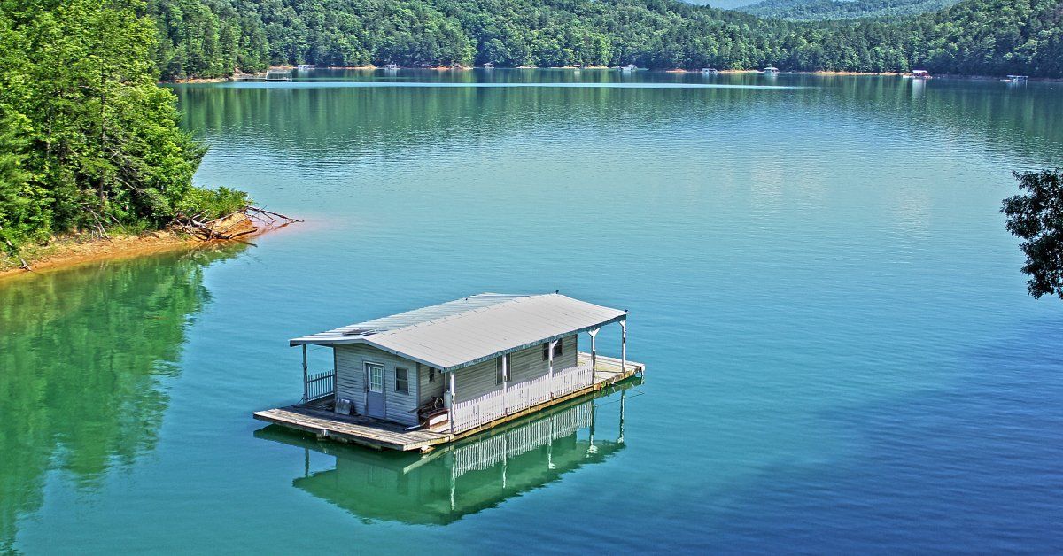 A gray house boat floating in the middle of a large body of water surrounded by mature trees.