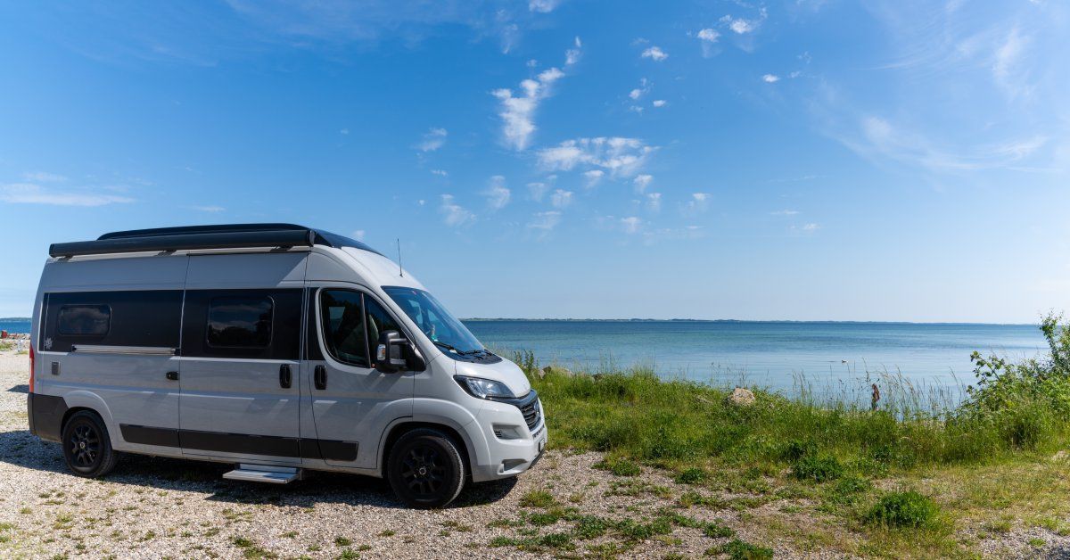 A gray and black camper van parked on gravel next to a body of water on a sunny day with a blue sky.