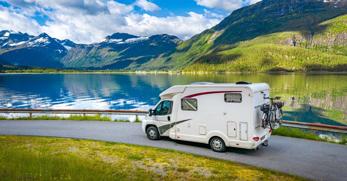 A white camper van with bikes attached to the back driving on a road next to a body of water and several mountains.