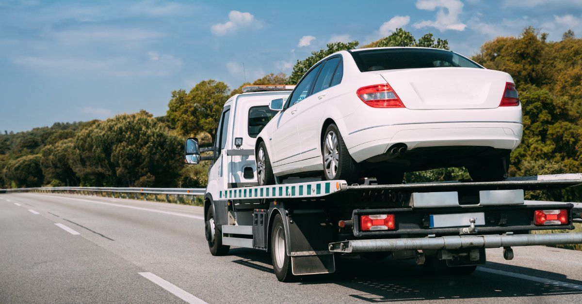 A tow truck with a white cab transporting a white sedan down a tree-lined road on a sunny day.