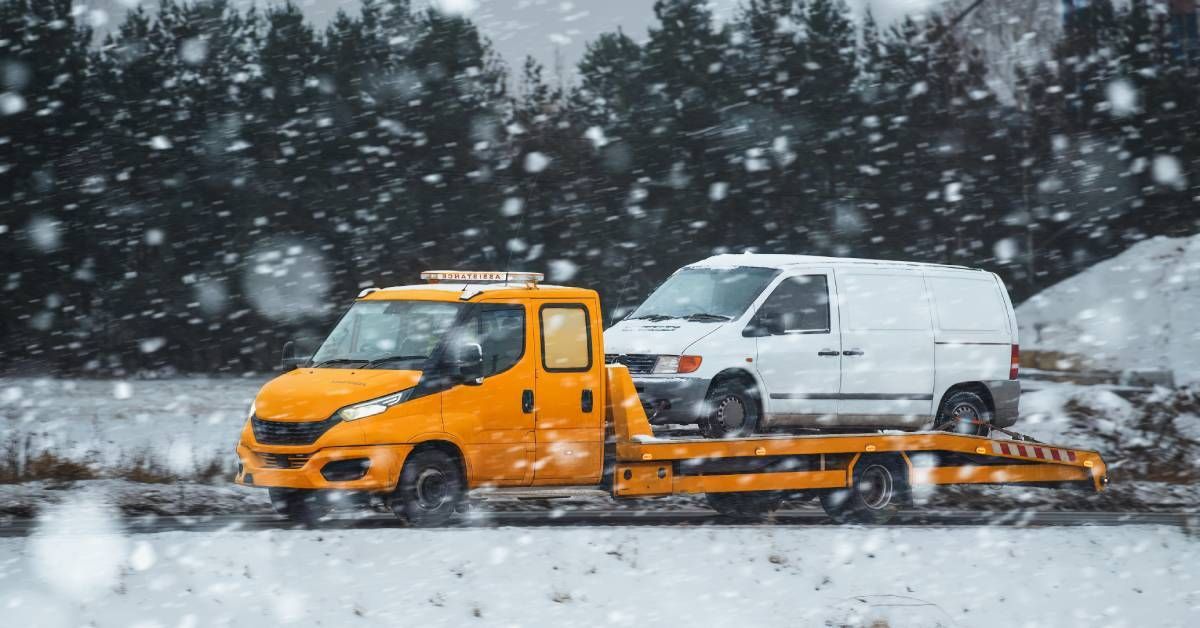 A truck with an orange cab and trailer transporting a white van on a snowy day with evergreen trees