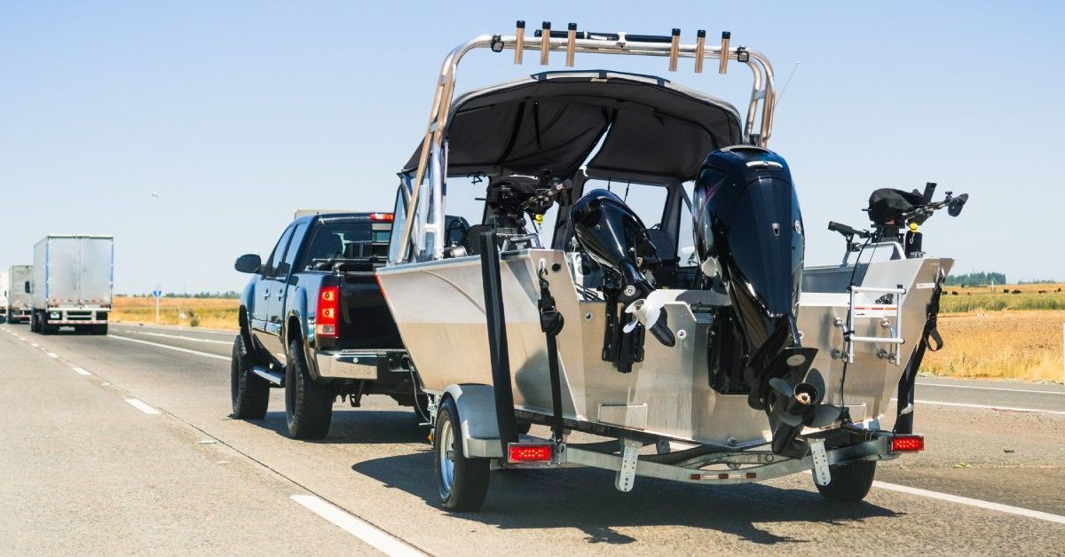 A black truck towing a silver and black motorboat on a trailer with two semi-trucks in the distance.