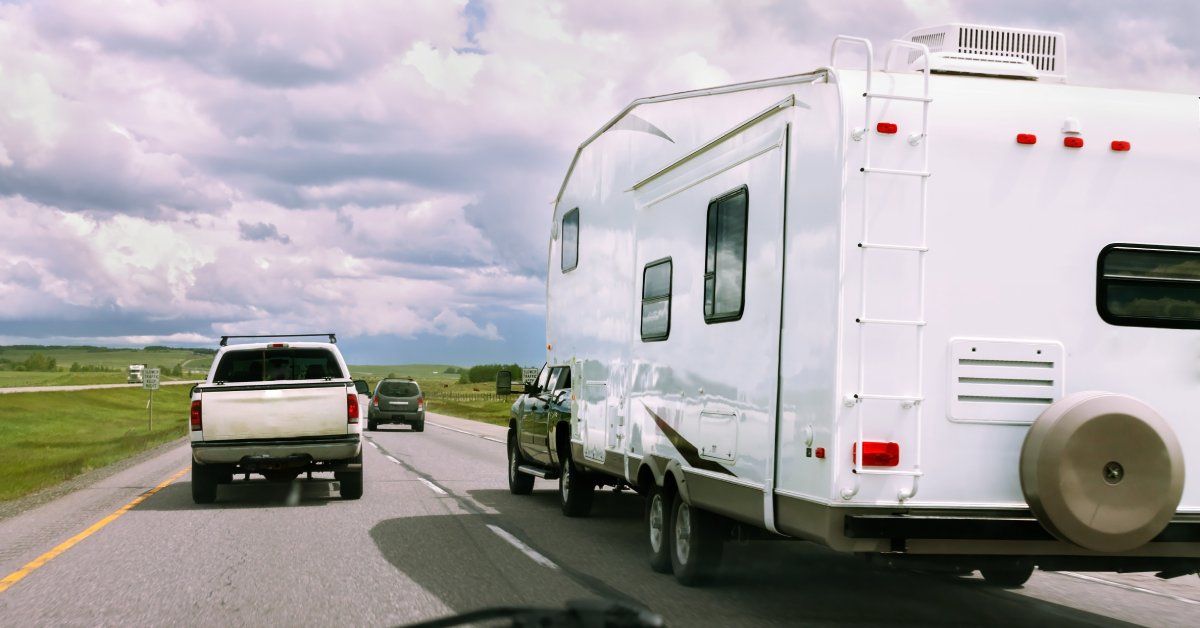 A black truck towing a large white RV travels on a divided highway, surrounded by other vehicles on a cloudy day.