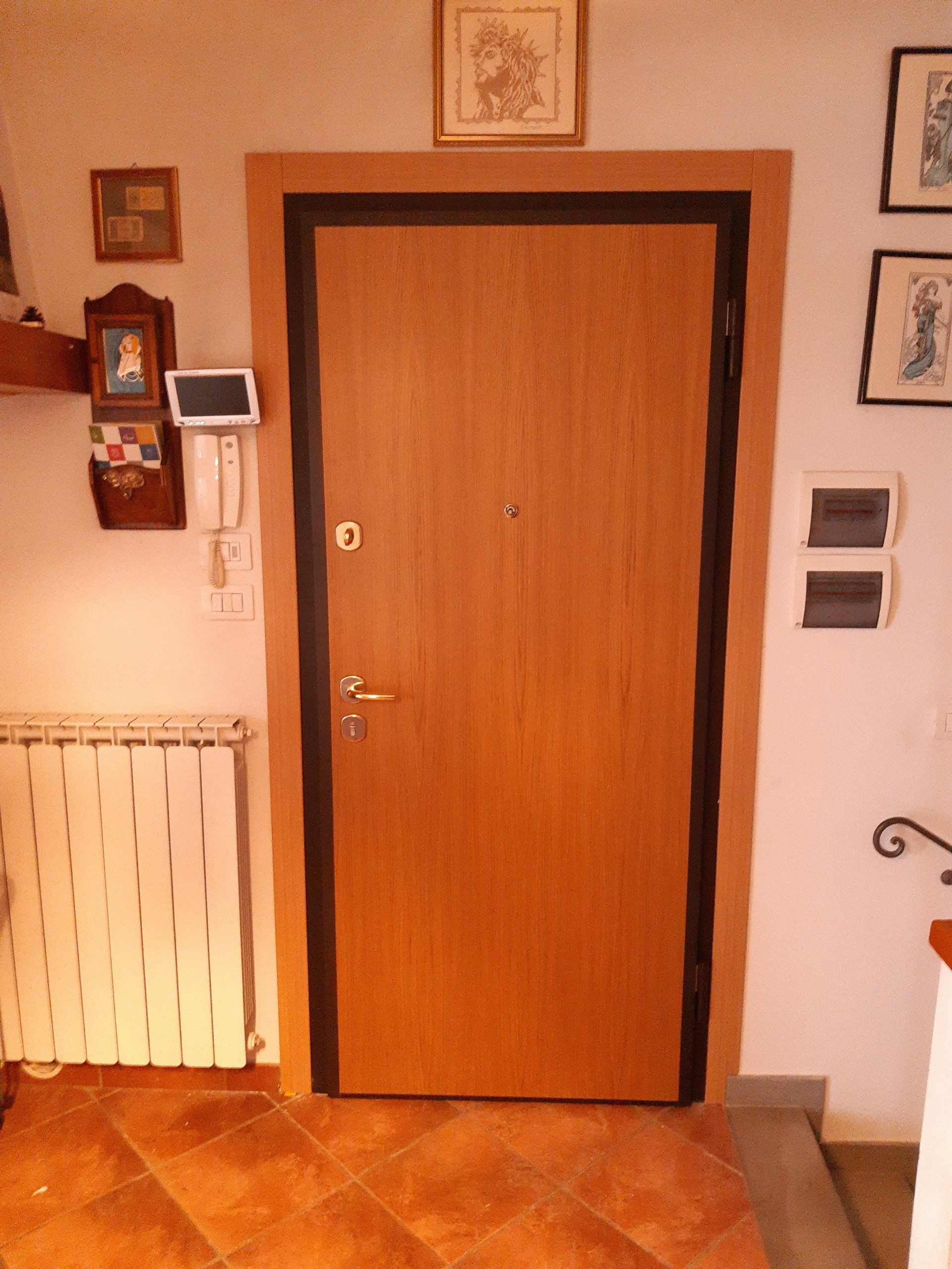 Porta d'ingresso in legno con cornici scure in un corridoio piastrellato, fiancheggiata da quadri incorniciati e un termosifone.