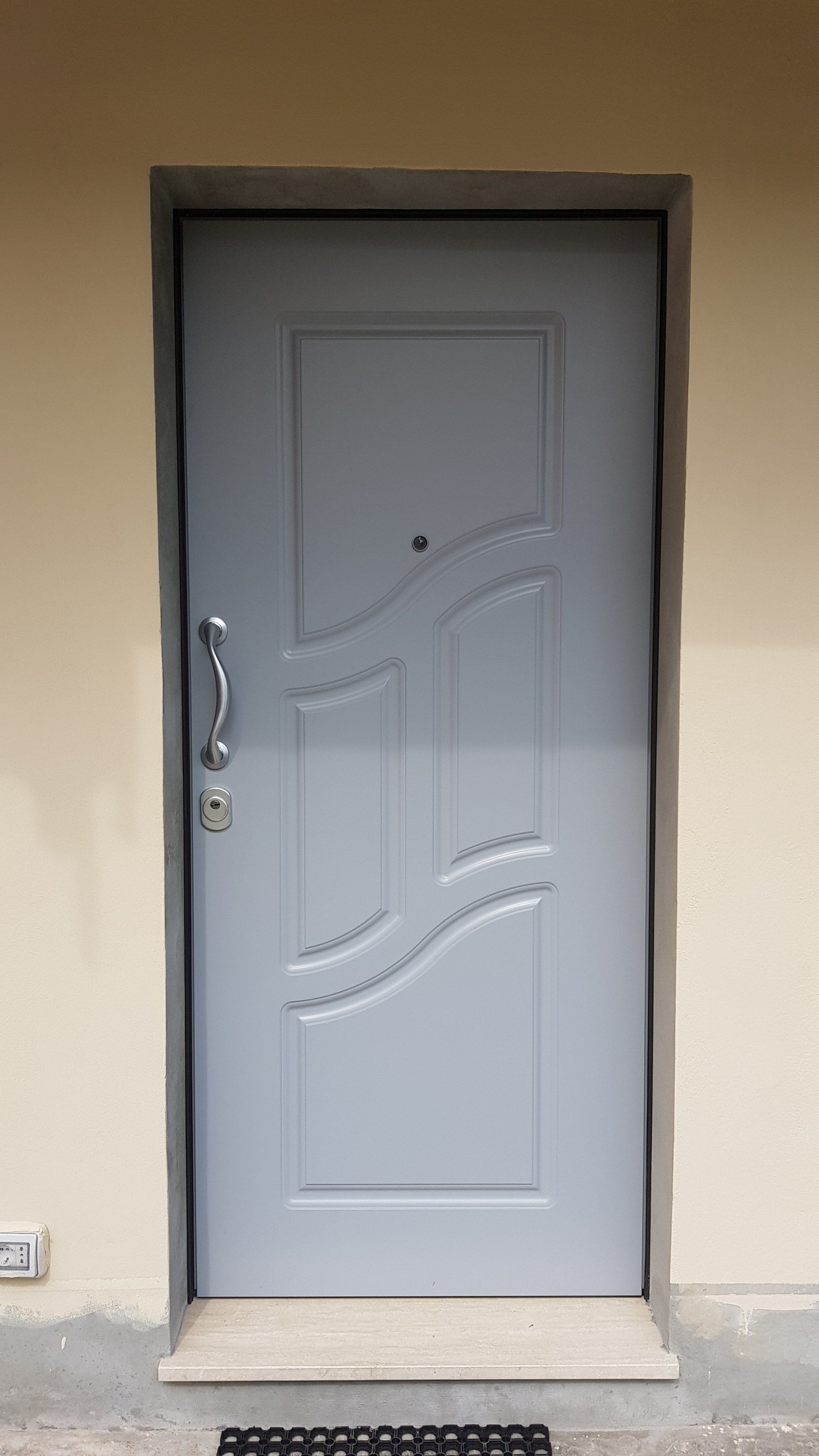 Porta d'ingresso grigia con pannelli decorativi ondulati incastonati in una parete beige.