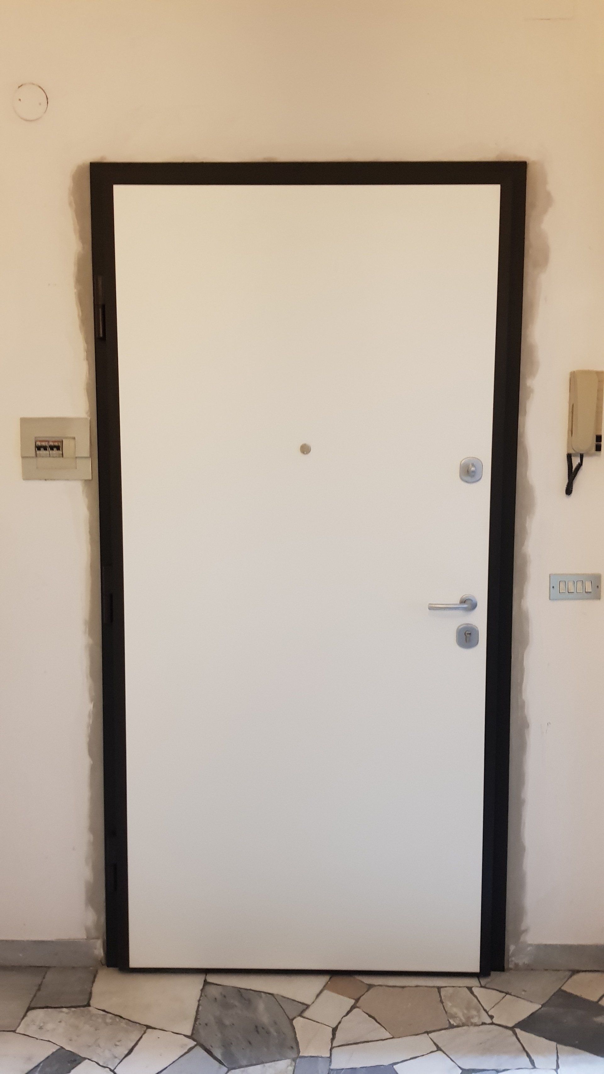 Porta d'ingresso bianca con finiture nere in un corridoio beige, con pavimento piastrellato a vista al di sotto.