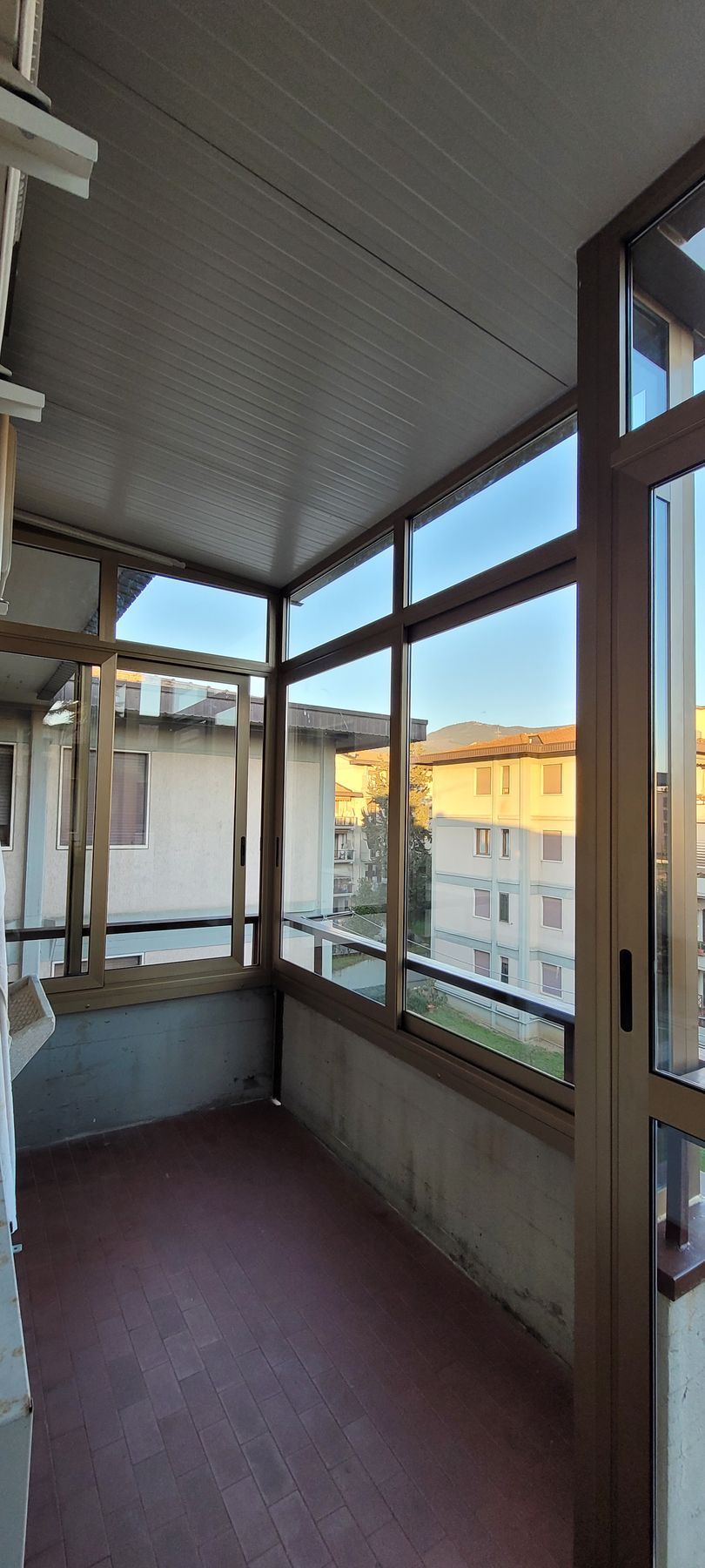 Balcone vuoto e chiuso, con ampie finestre e pavimento marrone, affacciato sugli edifici circostanti.