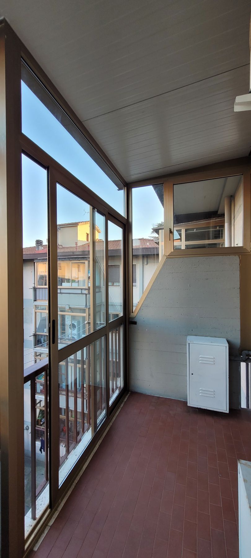 Stretto balcone di un appartamento con ringhiera in metallo, pavimento rosso ed elettrodomestico bianco, adiacente agli edifici vicini.