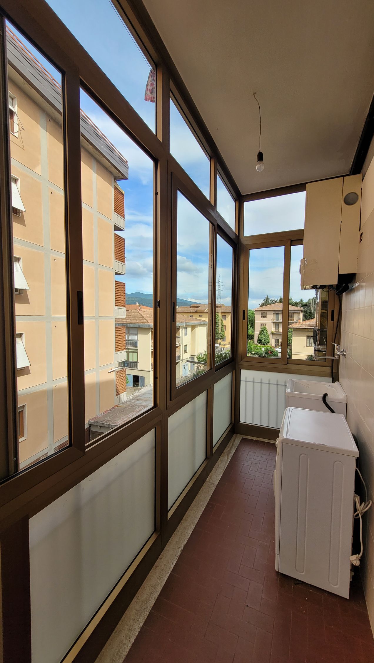Stretto balcone chiuso con ampie finestre, termosifoni e vista sui palazzi circostanti.