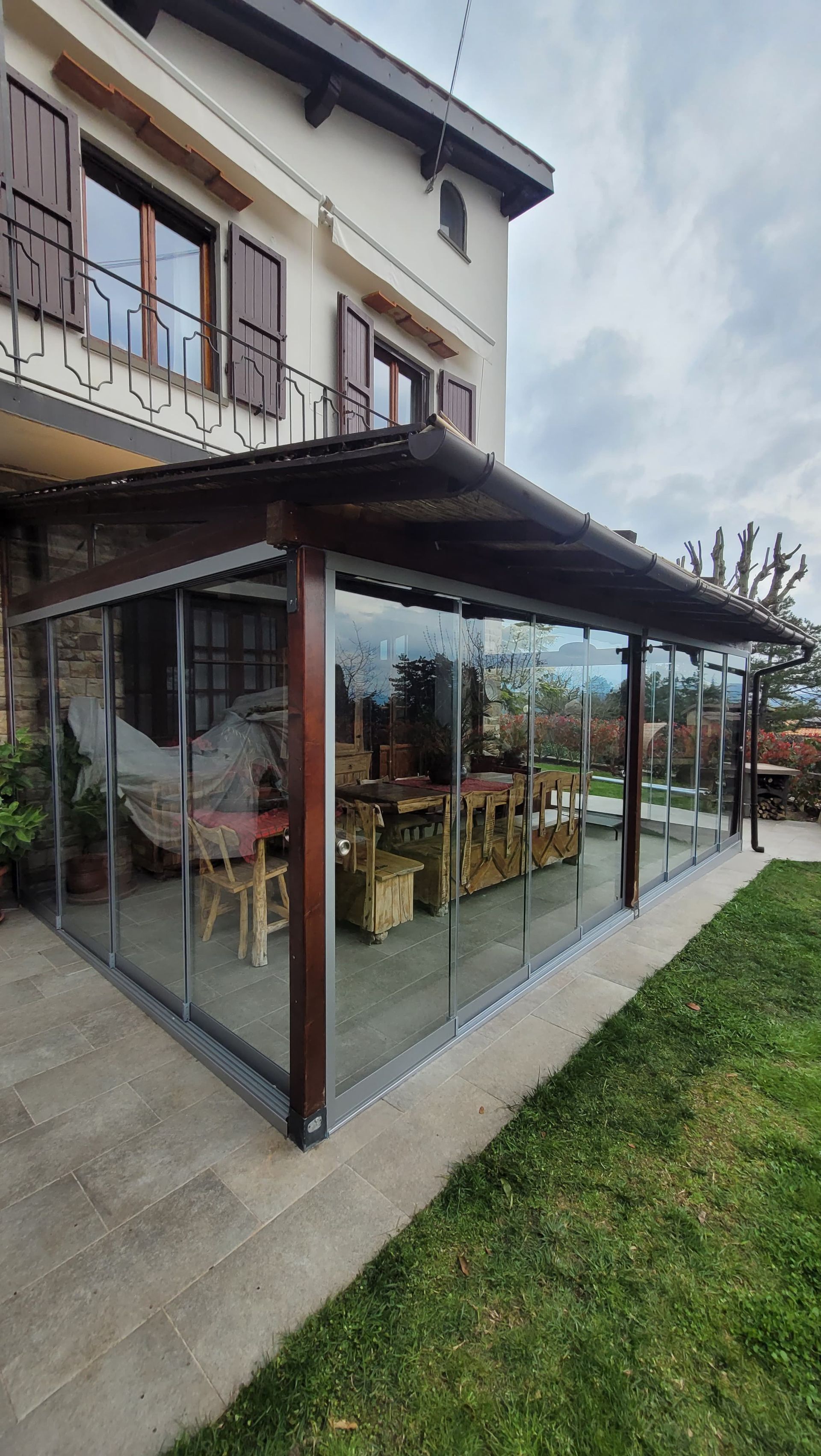 Patio con pareti in vetro adiacente a una casa, arredato con tavoli e sedie in legno sotto una tettoia scura.