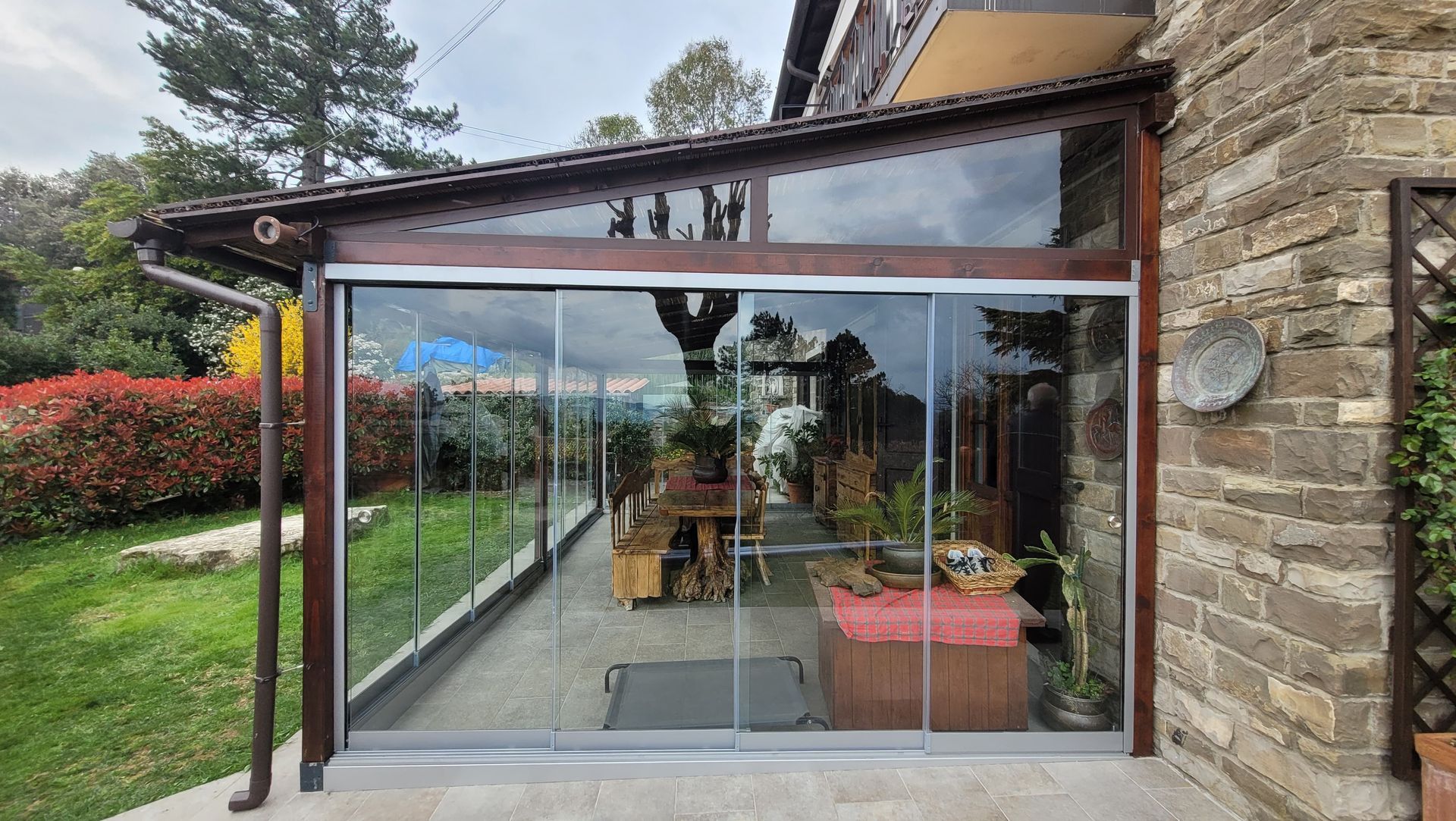 Veranda moderna in vetro annessa a una casa in pietra, con mobili da giardino e vista sul giardino.