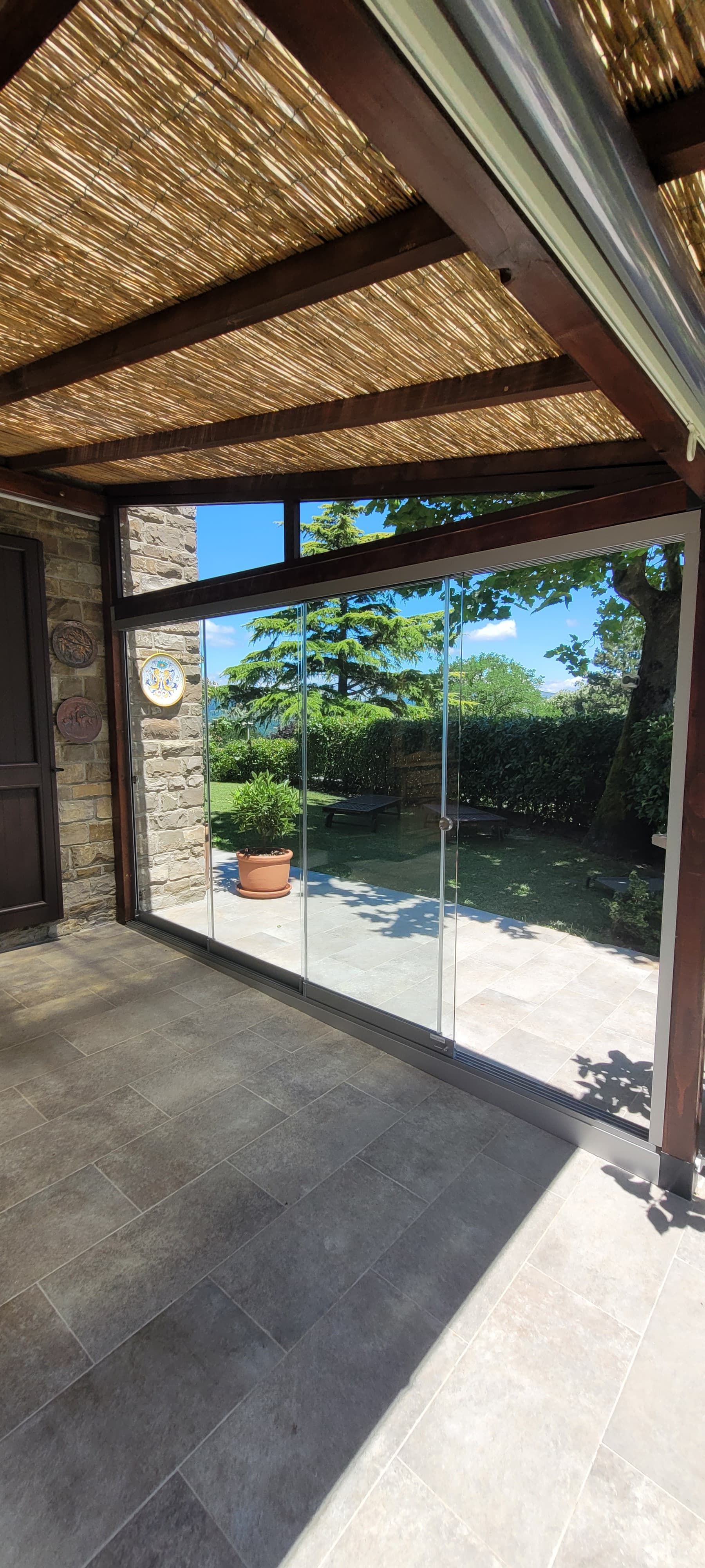 Patio coperto con soffitto in bambù, porte scorrevoli in vetro e una soleggiata vista sul giardino oltre la terrazza.