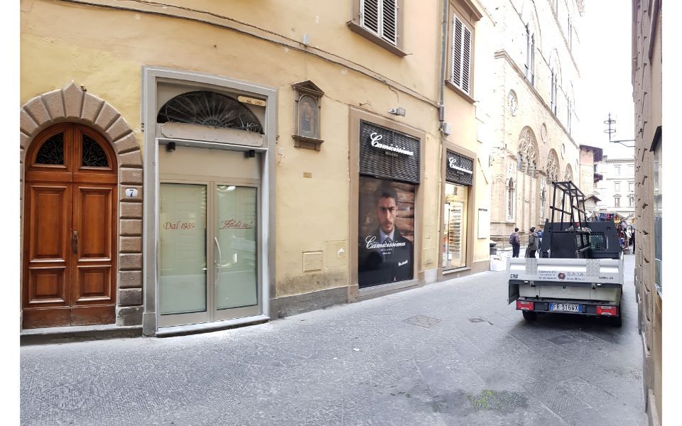 Stretta via europea con vetrine, uno scooter parcheggiato e un piccolo furgone sulla destra.