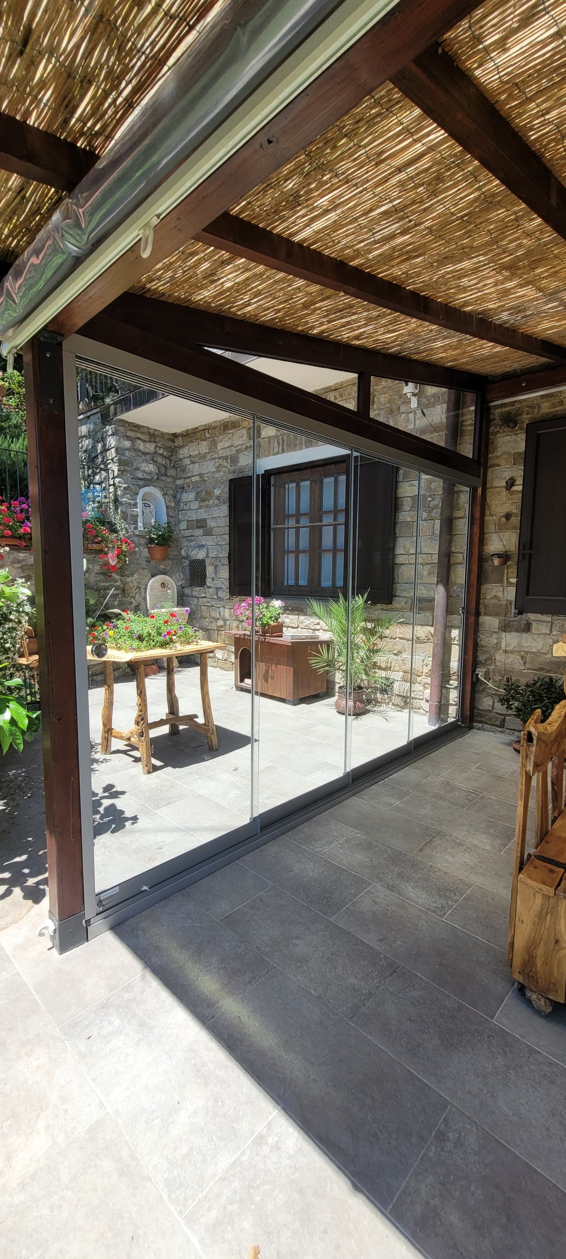 Patio coperto con pavimento in pietra, travi scure e piante in vaso, che si apre su un cortile con giardino.